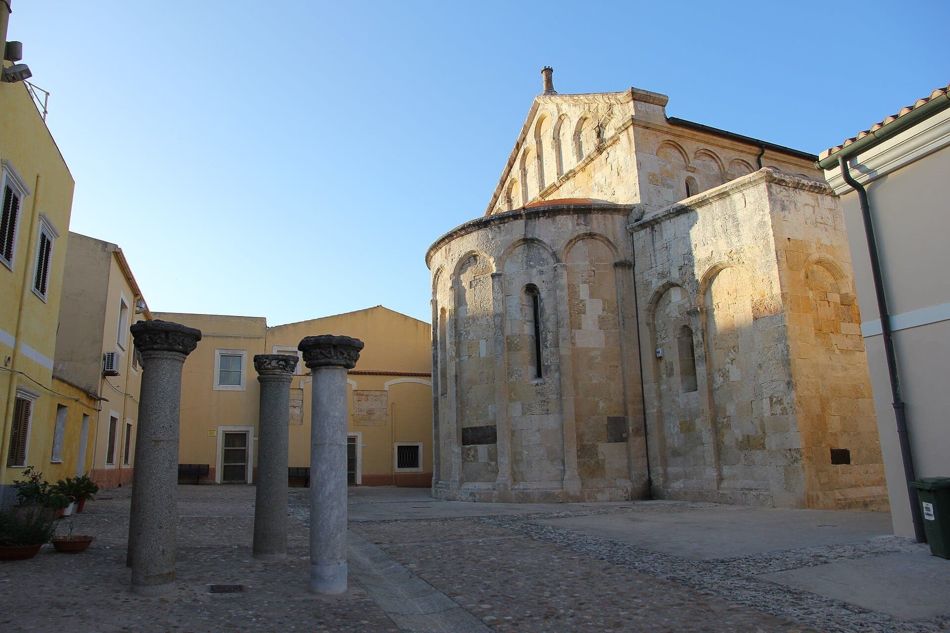 Columnas de piedra y el ábside redondeado de la Basilica di San Gavino en un patio soleado rodeado de edificios de color amarillo pastel en Porto Torres.