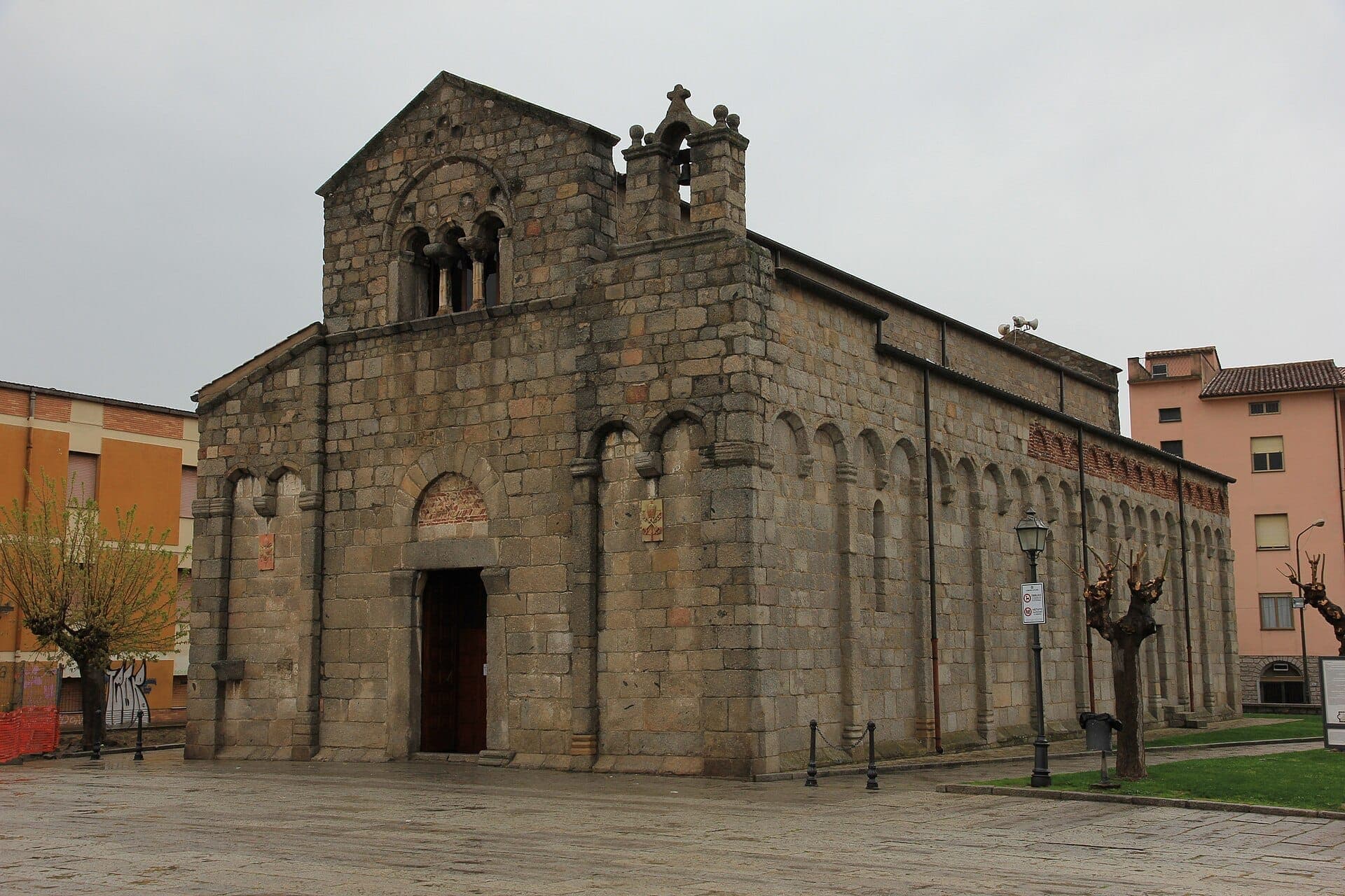 Vista frontal de la fachada de piedra y los arcos románicos de la Basilica di San Simplicio, rodeada de una plaza pavimentada y árboles recortados.