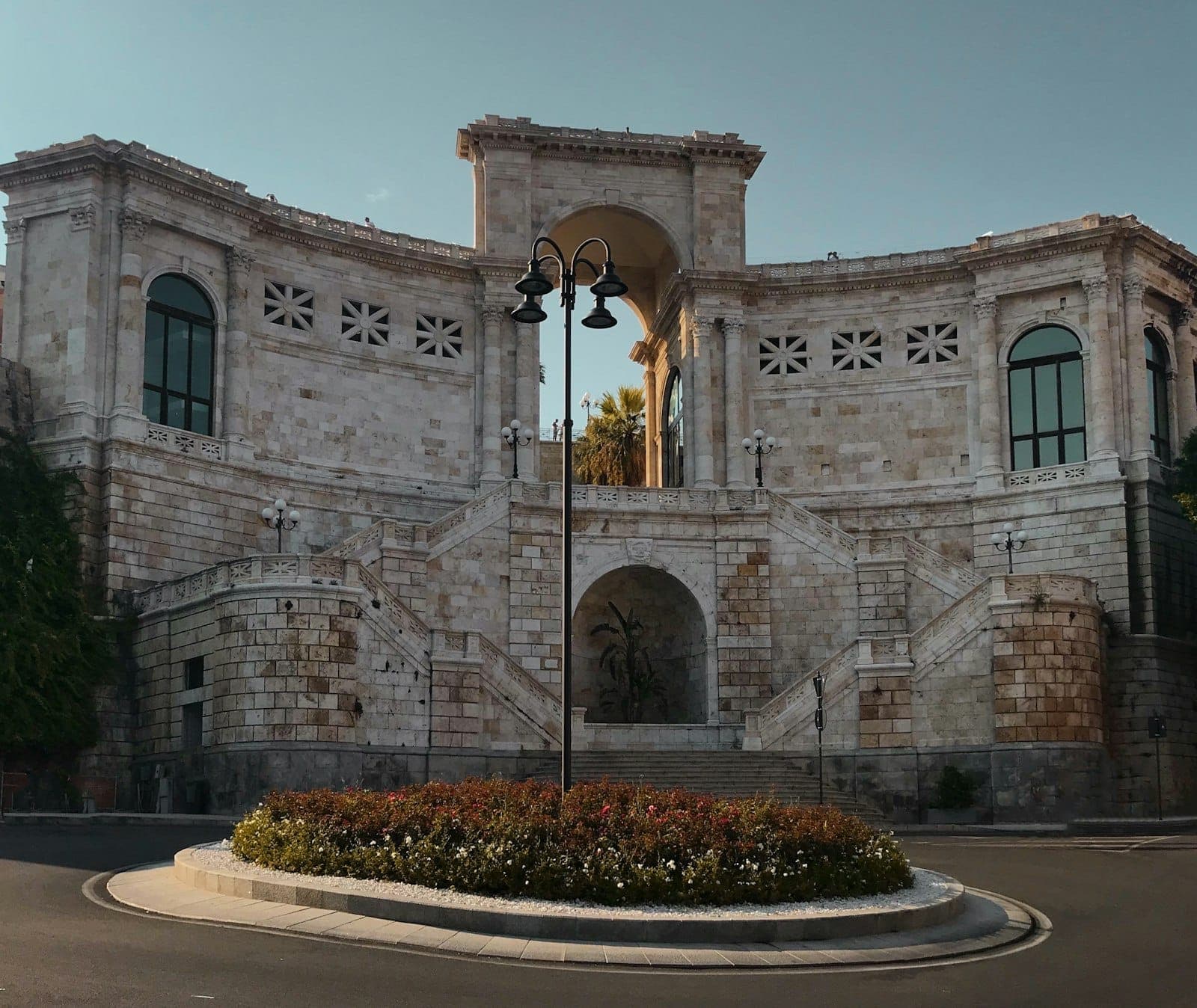 El Bastione di Saint Remy en Cagliari presenta grandes arcos de piedra blanca, escaleras dobles y una rotonda con flores bajo un cielo despejado.
