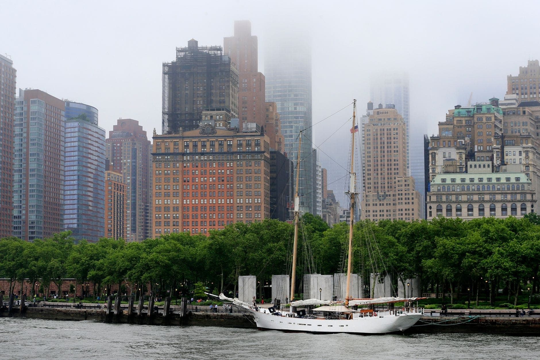 เรือใบขาวจอดอยู่ริมน้ำ Battery Park ท่ามกลางต้นไม้เขียวร่มรื่น อาคารเก่าแก่แมนฮัตตันโผล่ท่ามกลางหมอกจางๆ ด้านหลัง
