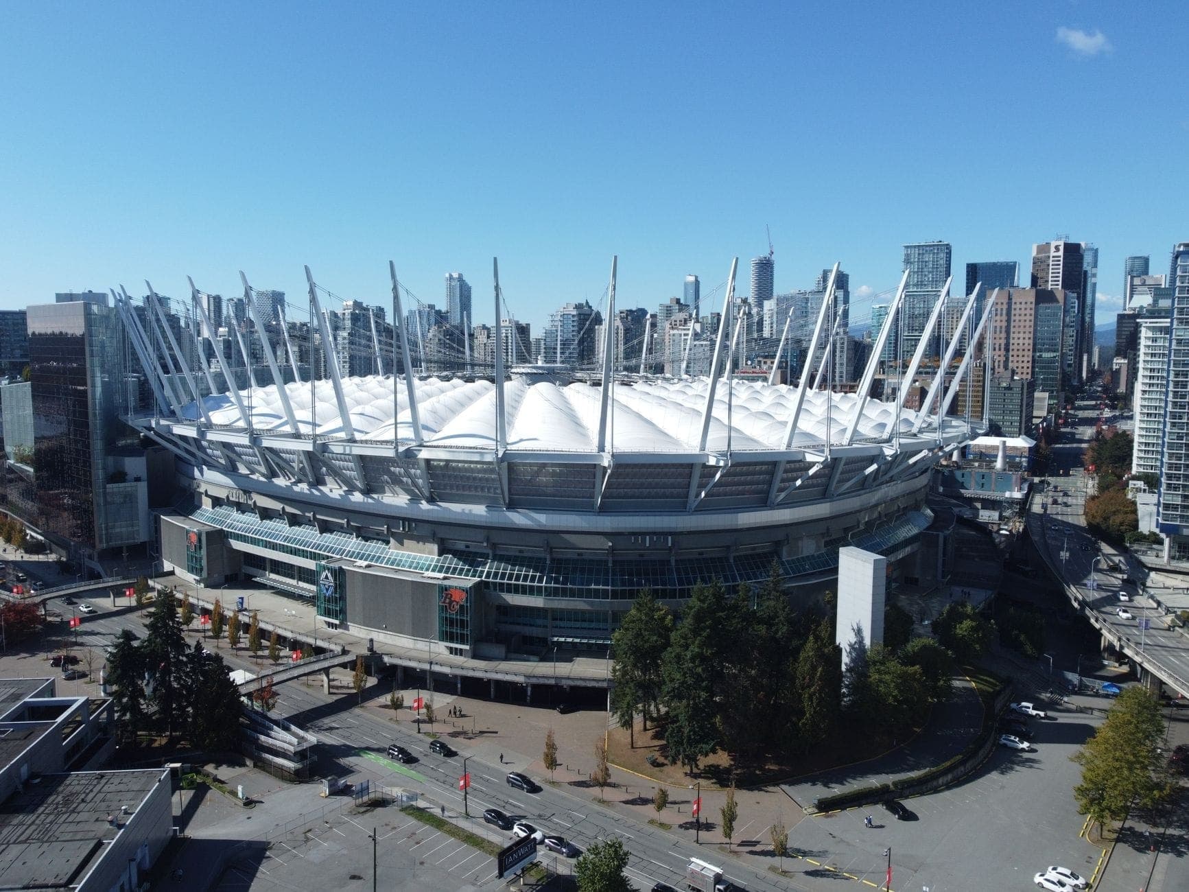 มุมมองทางอากาศของสนามกีฬา BC Place พร้อมหลังคาพับเก็บได้สีขาวอันโดดเด่น ล้อมรอบด้วยตึกระฟ้าสมัยใหม่ของ Vancouver ภายใต้ท้องฟ้าสีฟ้าใส
