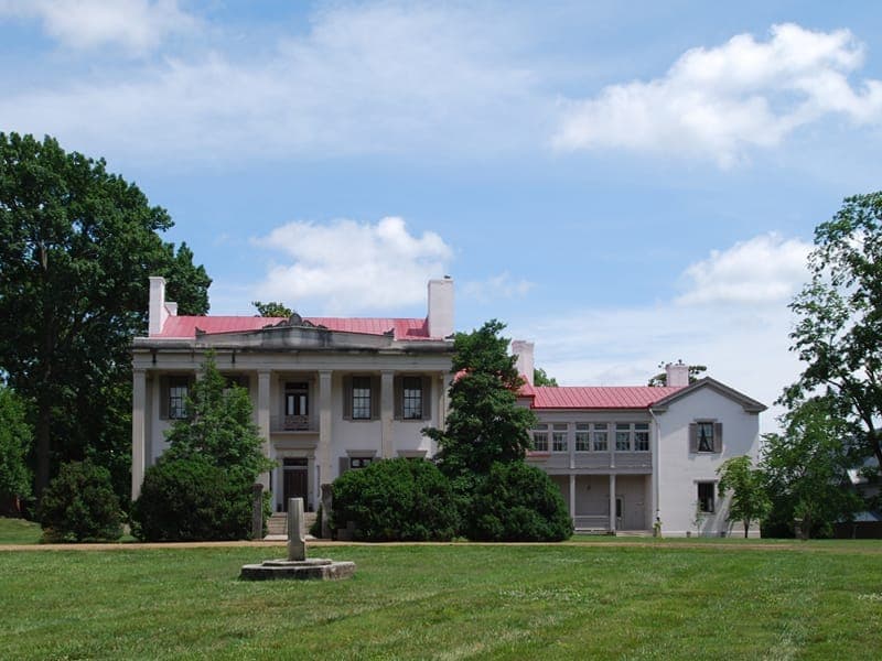 Belle Meade Plantation mansion with white columns and red roof, surrounded by green lawns, bushes, and tall trees on a sunny day.