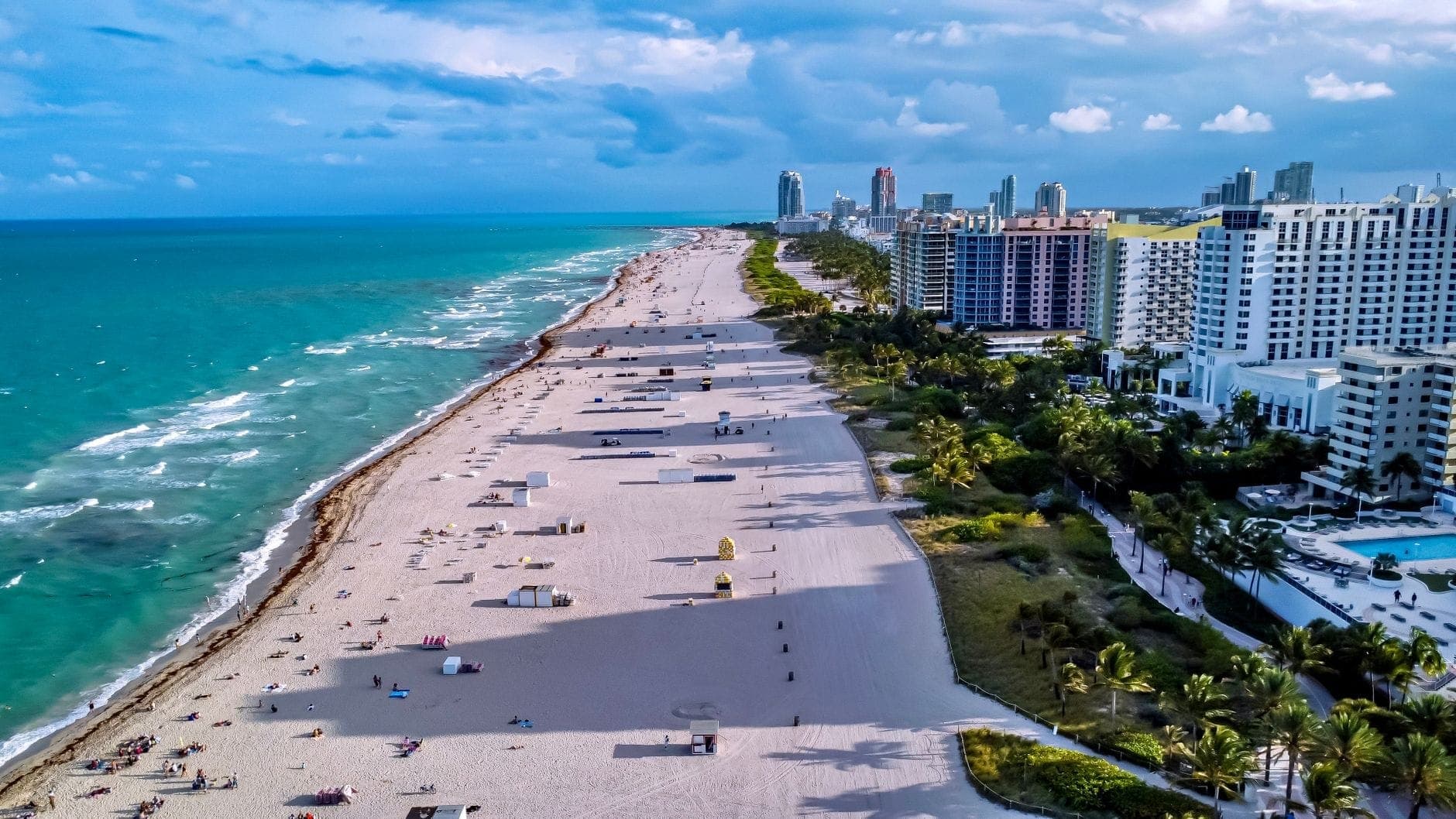 Aerial view of wide sandy Miami beach with scattered people, colorful lifeguard towers, and seaside hotels lining the Upper Miami Beach Strip.
