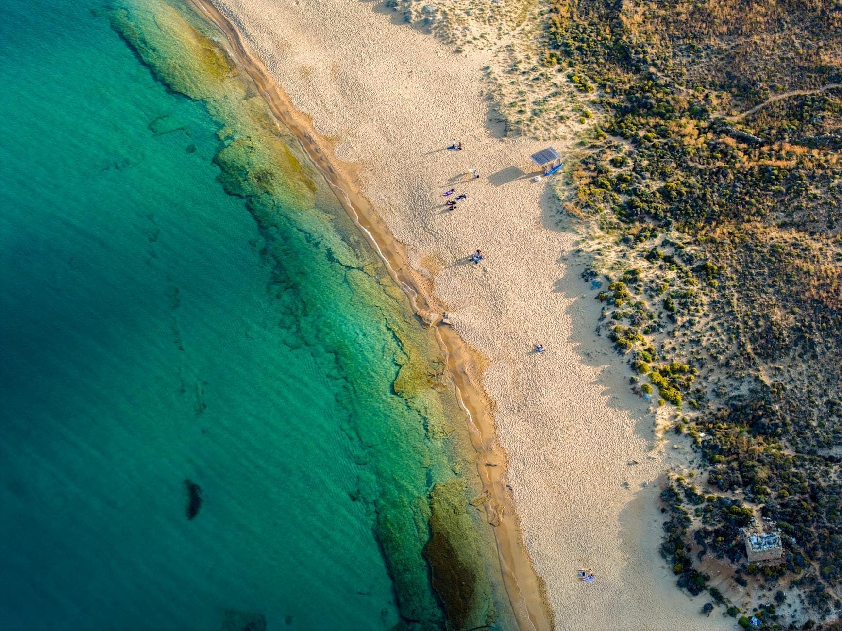 Luftaufnahme eines ruhigen, unbebauten Sandstrandes mit klarem türkisfarbenem Wasser und vereinzelten Besuchern, umgeben von natürlicher Landschaft ohne Gebäude.