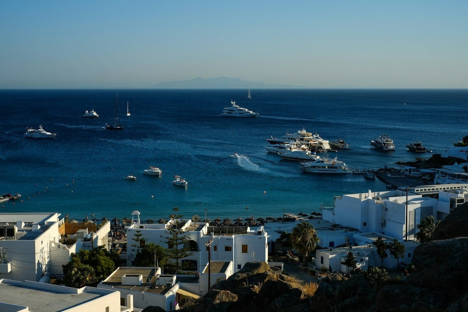 Yachten und Boote vor Anker nahe einem Sandstrand in Mykonos, mit weißen Gebäuden und blauem Meer unter klarem Himmel.