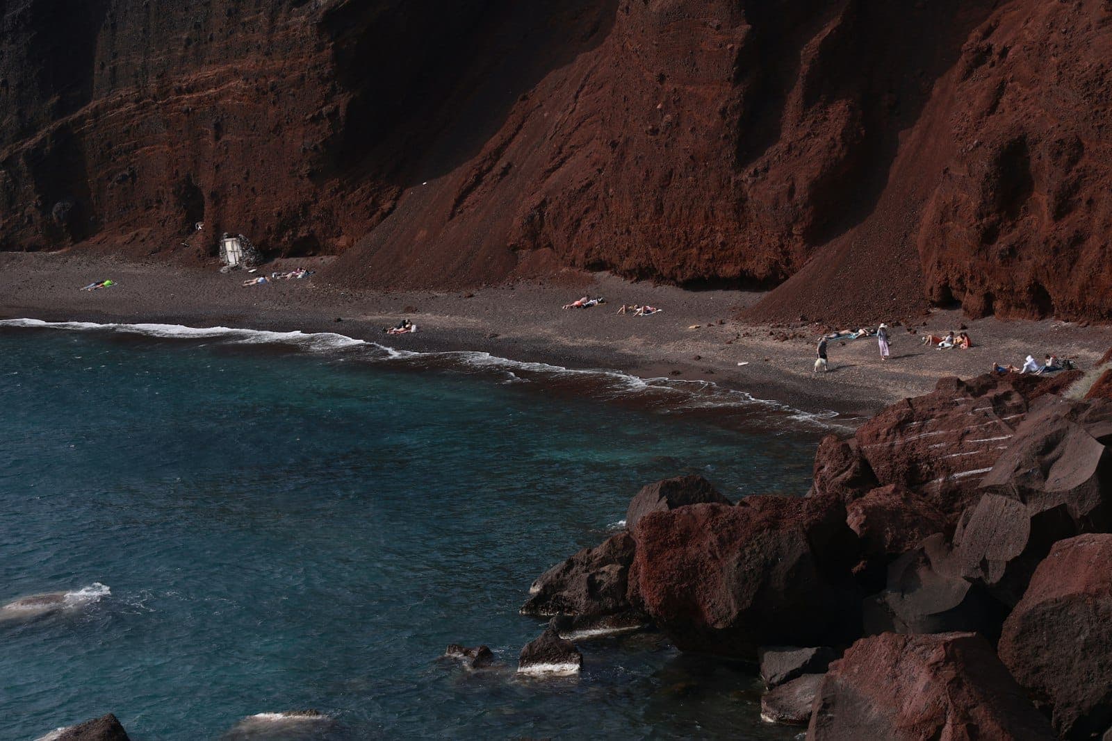 Dramatischer Blick auf den Roten Strand Santorins mit markanten roten Vulkanfelsen, schwarzem Sand und tiefblauem Wasser, eingerahmt von Felsvorsprüngen und Sonnenanbetern am Ufer.