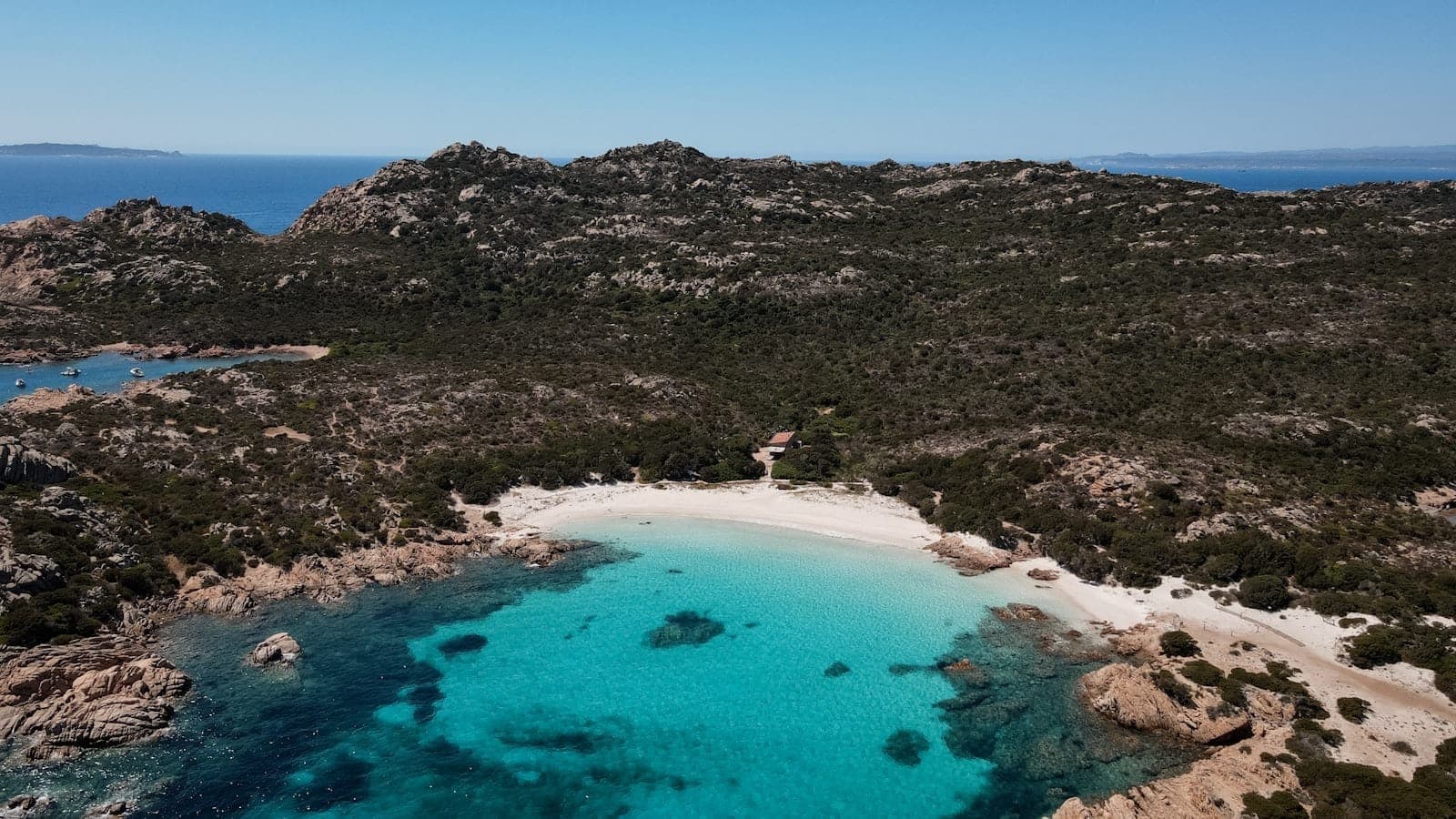Luftaufnahme einer kleinen Bucht mit türkisfarbenem Wasser, weißem Sandstrand und beeindruckenden Granitfelsen, umgeben von mediterranem Buschland.