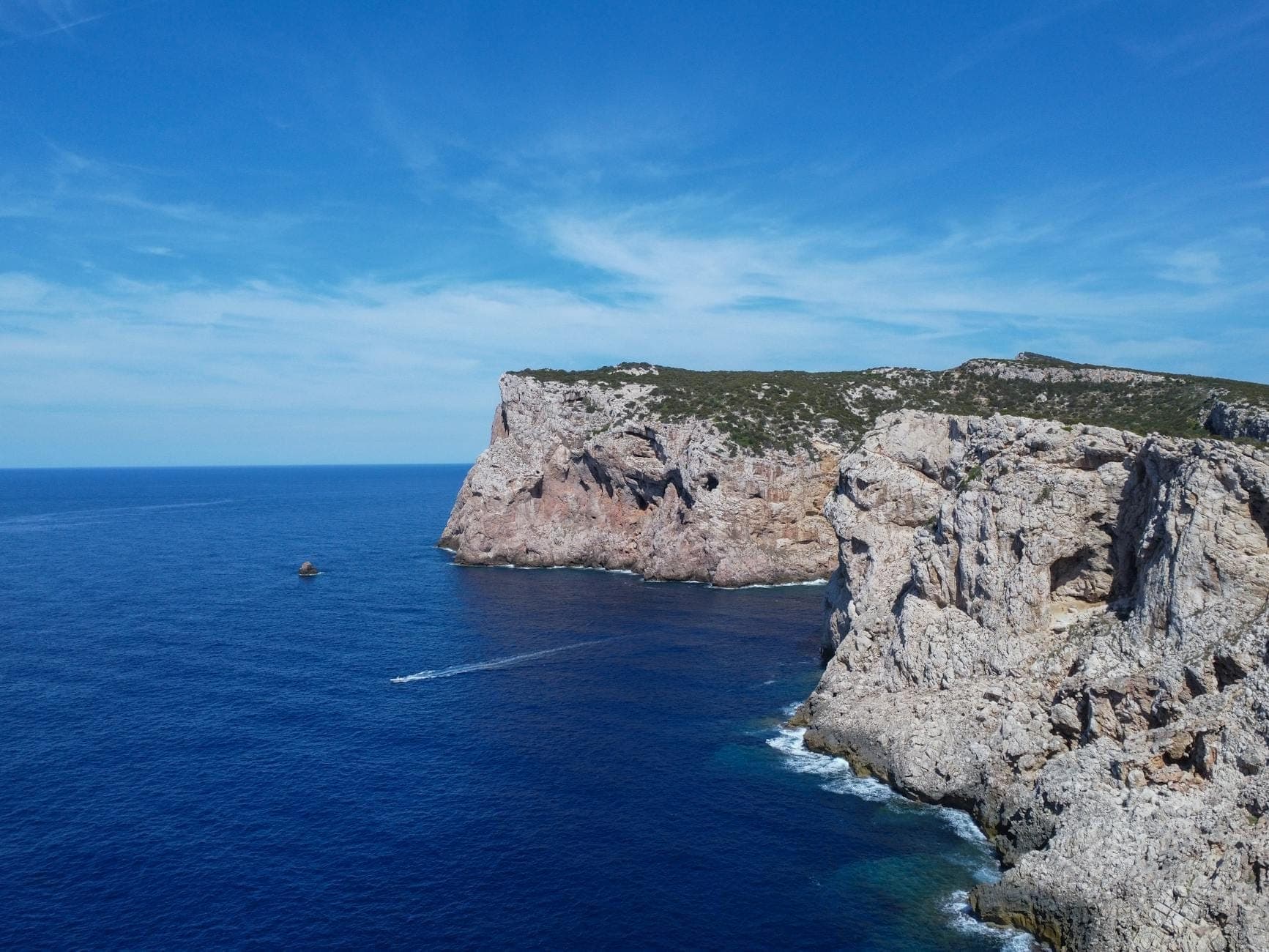 Dramatische Klippen mit Blick auf das tiefblaue Meer mit einem Boot und einer zerklüfteten Küste im Nordwesten Sardiniens unter klarem Himmel.