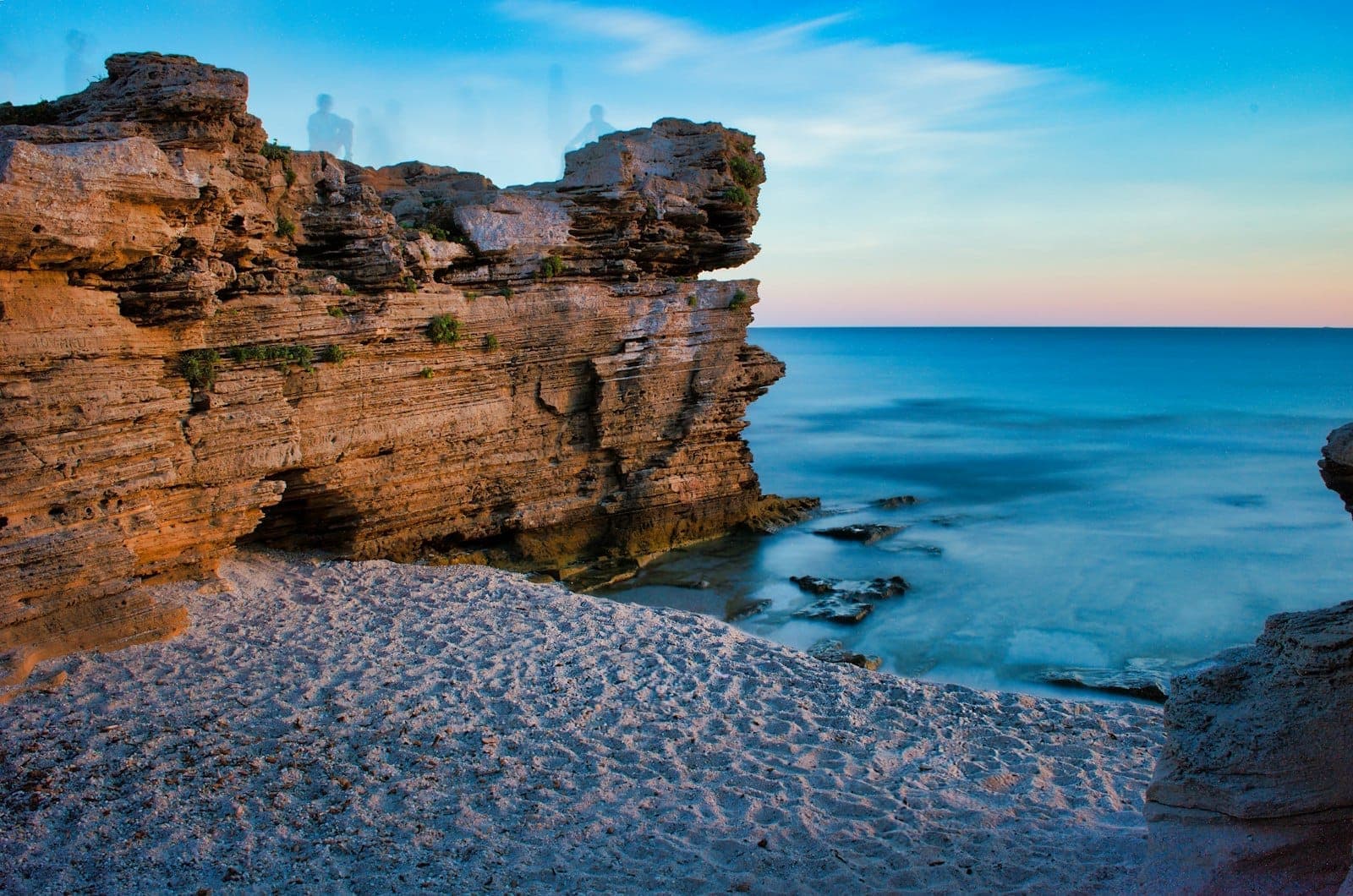 Felsige Meeressäulen mit geschichteten Klippen, die über einer kleinen Sandbucht aufragen, mit ruhigem blauem Wasser bei Sonnenuntergang.