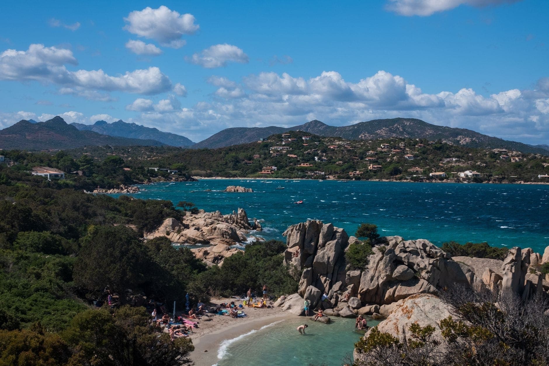 Ein malerischer Strand an der Costa Smeralda mit türkisblauem Wasser, markanten Granitfelsen, üppigem Grün und Menschen, die sich unter blauem Himmel sonnen.