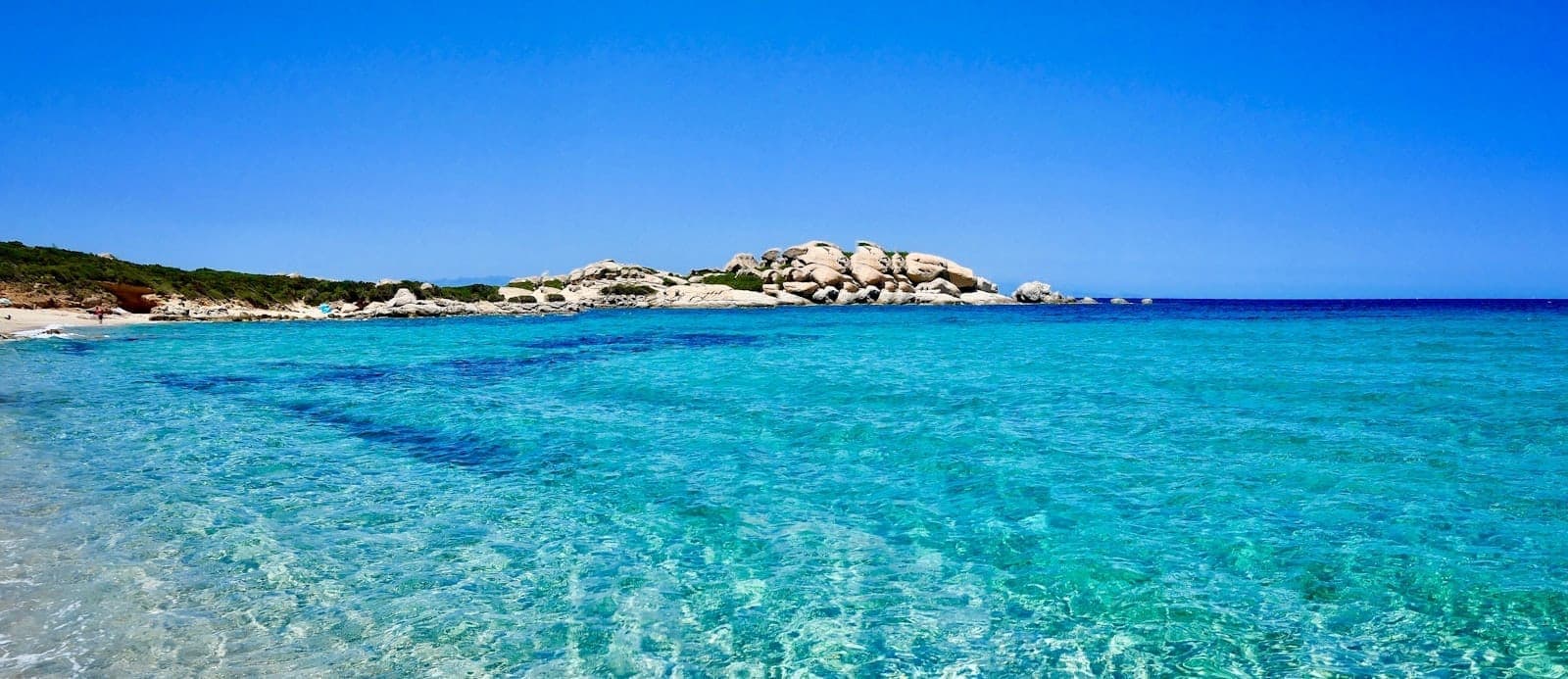 Weitläufige Landschaft eines sardischen Strandes mit kristallklarem türkisfarbenem Wasser, weißem Sand und felsigen Hügeln unter strahlend blauem Himmel – perfektes Titelbild für einen Reiseführer.