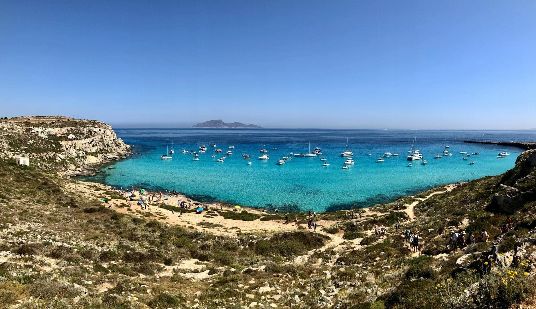 Panoramikong tingin ng isang kalansay na look sa mga isla ng Sicily na may kulay-turkesang tubig, mga bangkang nakaangkla, buhangin na dalampasigan, at mabatong baybayin sa ilalim ng malinaw na asul na langit.