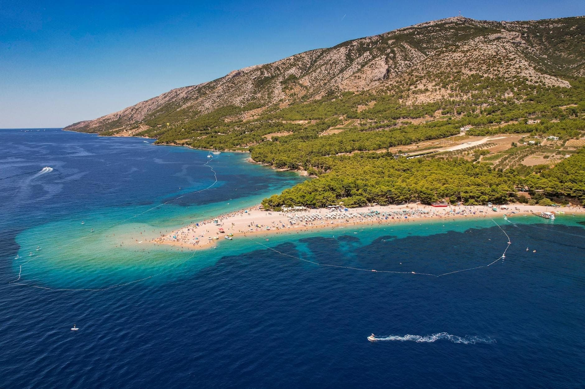 Vue aérienne de la plage Zlatni Rat sur l'île de Brač, avec eau turquoise, forêt de pins, flèche de sable et montagnes lointaines sous un ciel bleu.