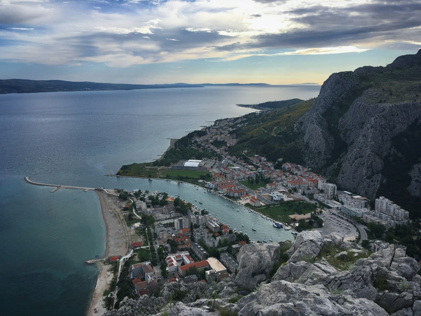 Ville côtière avec un mélange de plages de sable et de zones de galets, entourée de montagnes et d'un port de plaisance visible, probablement sur la côte dalmate.