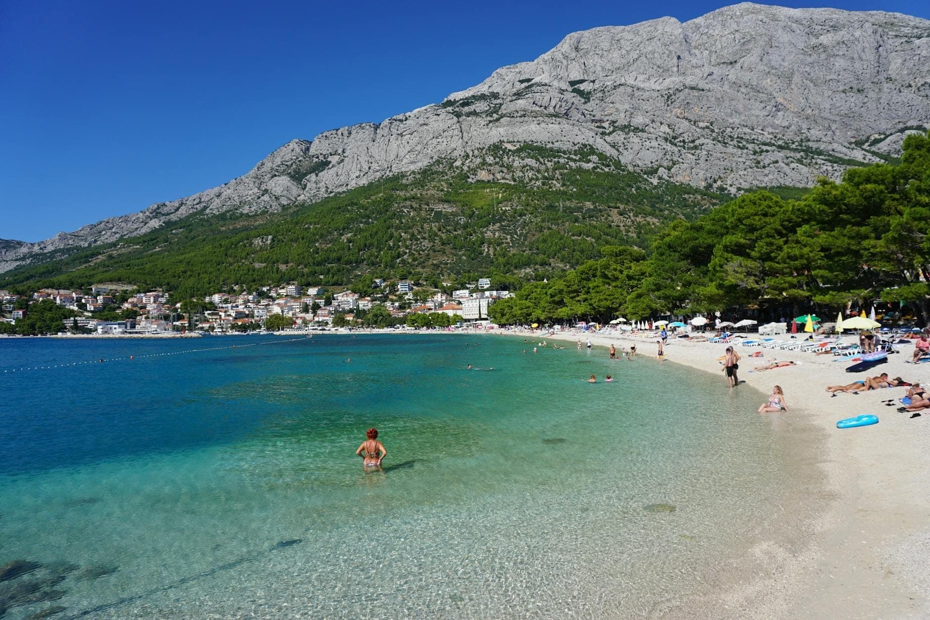 Des baigneurs profitent d'une plage de galets aux eaux turquoise près de Split, entourée d'arbres verts et de montagnes rocheuses en arrière-plan.