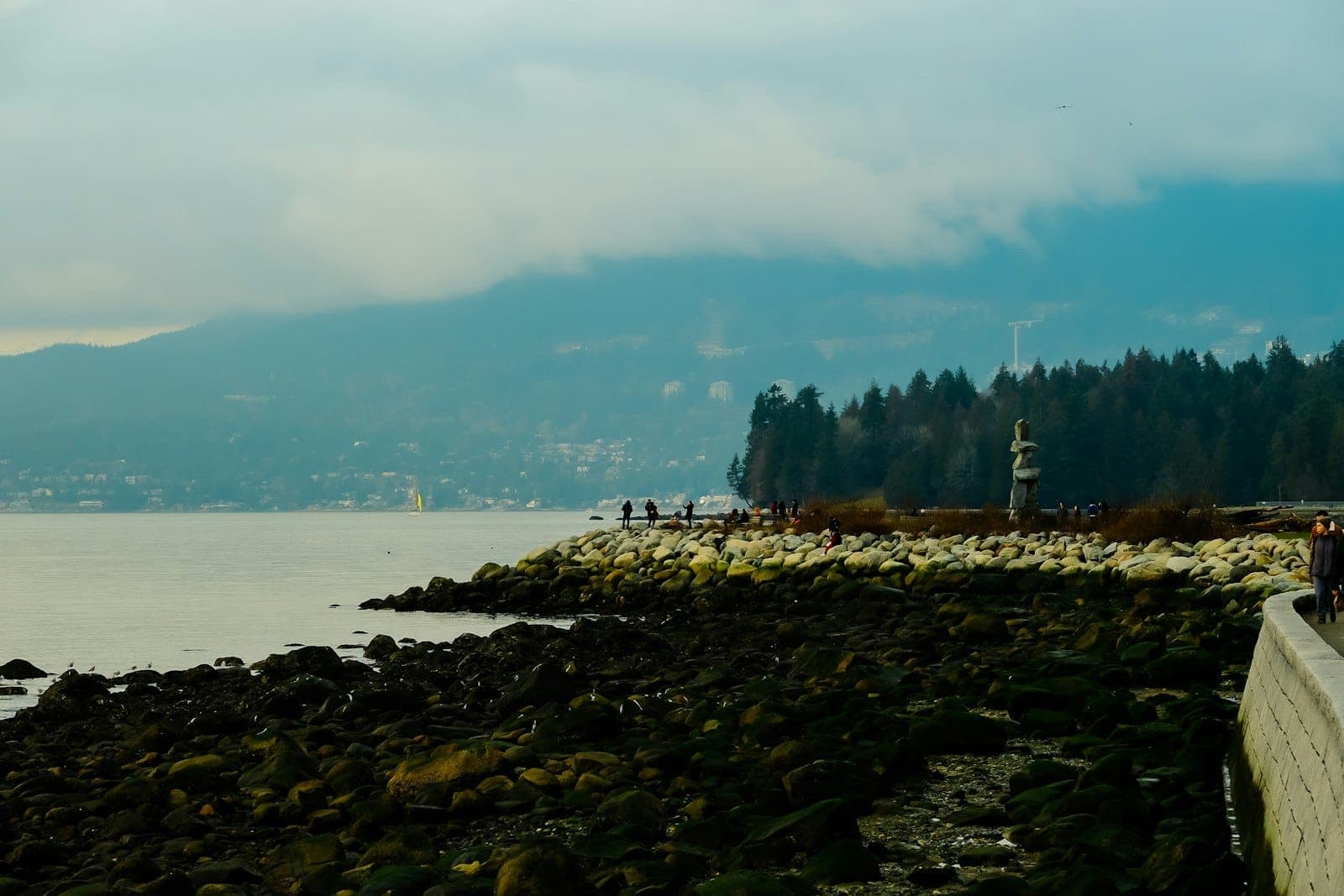 วิวทางเดินริมทะเลของ Stanley Park พร้อมรูปปั้น Inukshuk ชายฝั่งหิน และภูเขาของ Vancouver เป็นฉากหลัง