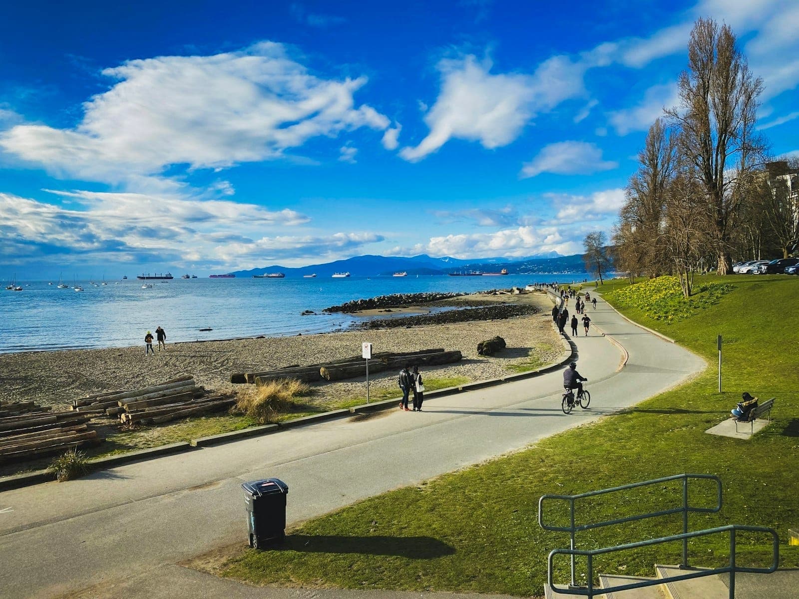 Vancouver Seawall ที่คึกคักด้วยผู้คนปั่นจักรยานและเดินเล่นริมชายหาด ท่อนซุง มหาสมุทร และวิวภูเขาอันกว้างไกลใต้ท้องฟ้าสดใส