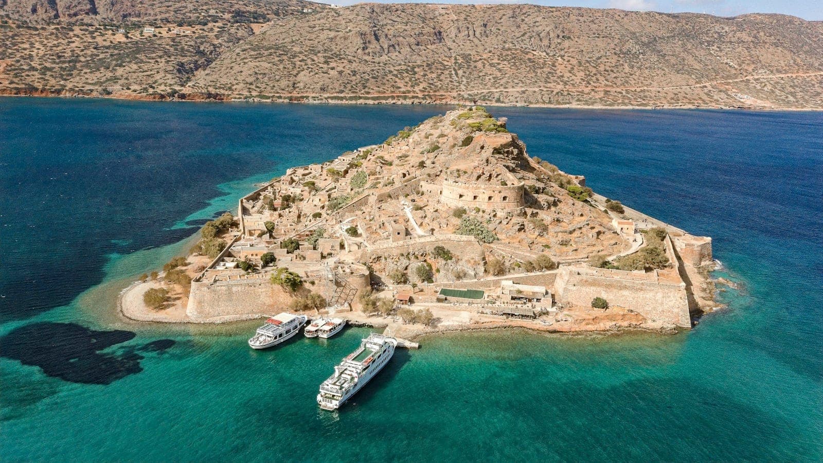 Luftaufnahme der Festung der Insel Spinalonga vor der Küste mit türkisfarbenem Meer und angedockten Ausflugsbooten, bergige Landschaft im Hintergrund.
