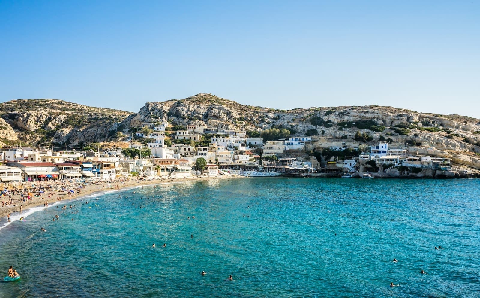 Sonniges Küstendorf auf Kreta mit türkisfarbenem Wasser, Badenden, Sandstrand, weiß getünchten Häusern und felsigen Hügeln unter strahlend blauem Himmel.