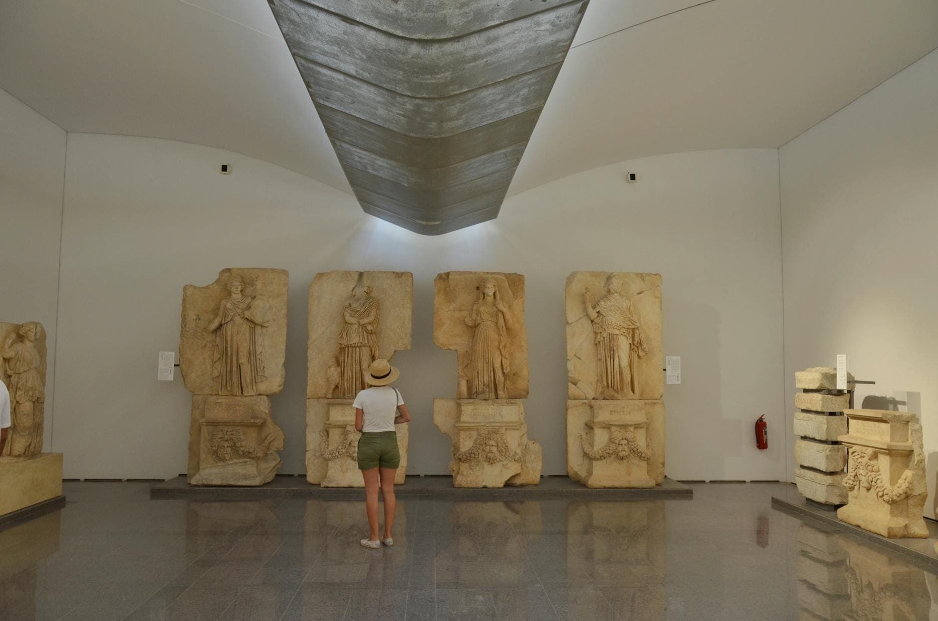 Seseorang berdiri di museum arkeologi modern, mengamati relief batu kuno berukuran besar di galeri yang terang.
