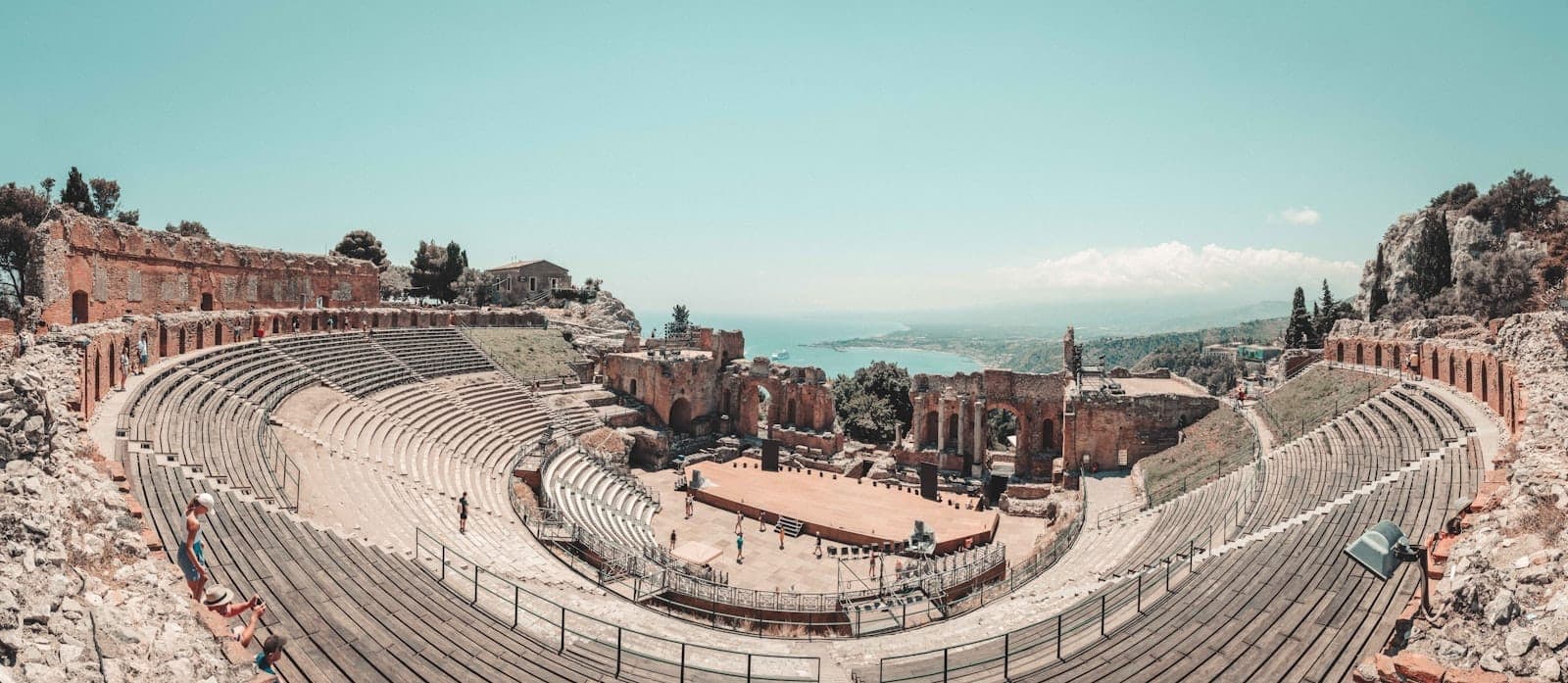 Pemandangan panoramik luas teater Yunani kuno di Taormina, Sisilia, dengan tempat duduk batu, reruntuhan panggung, dan garis pantai yang terlihat di latar belakang.
