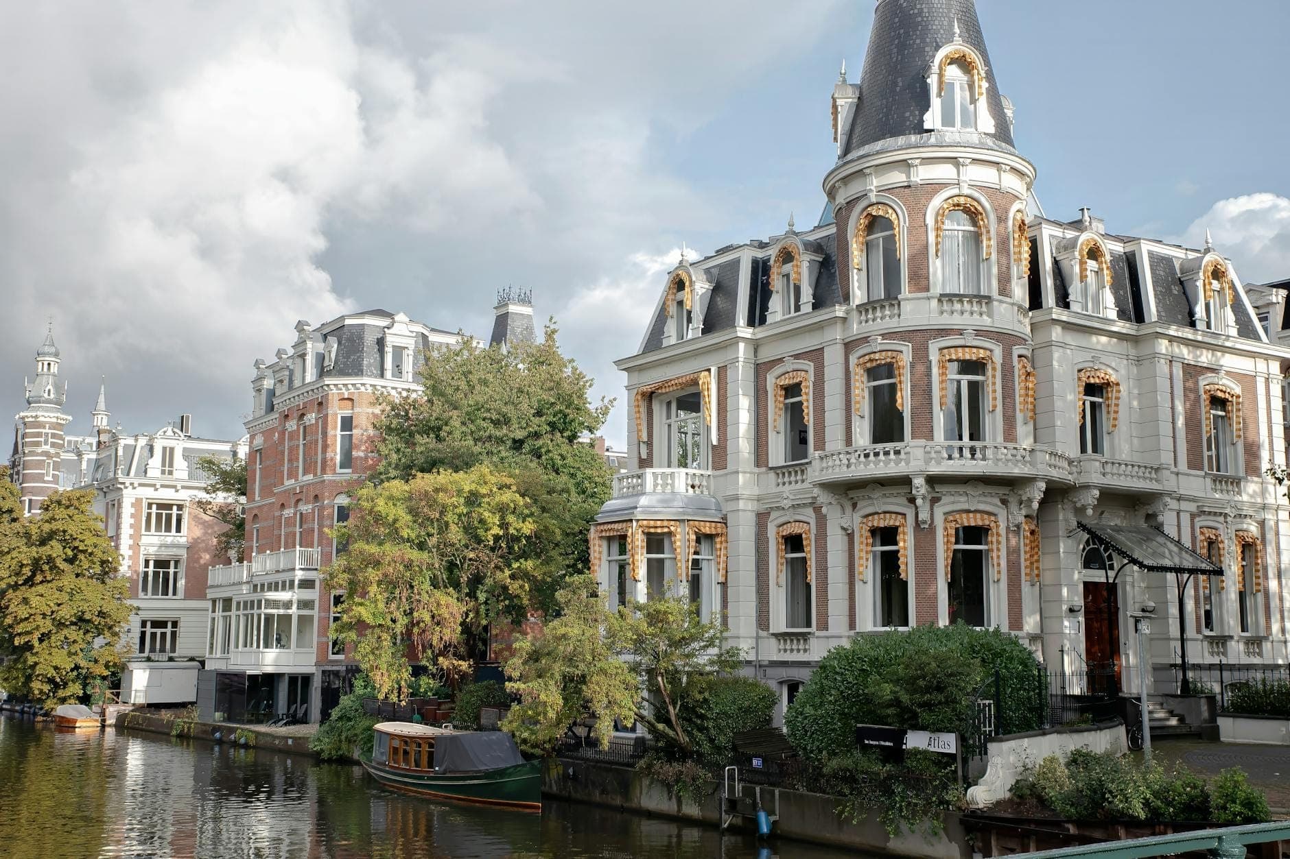 Verziertes historisches Herrenhaus an einem Kanal in Amsterdam, mit üppigem Grün, klassischer Architektur und Kanalbooten unter einem teils bewölkten Himmel.