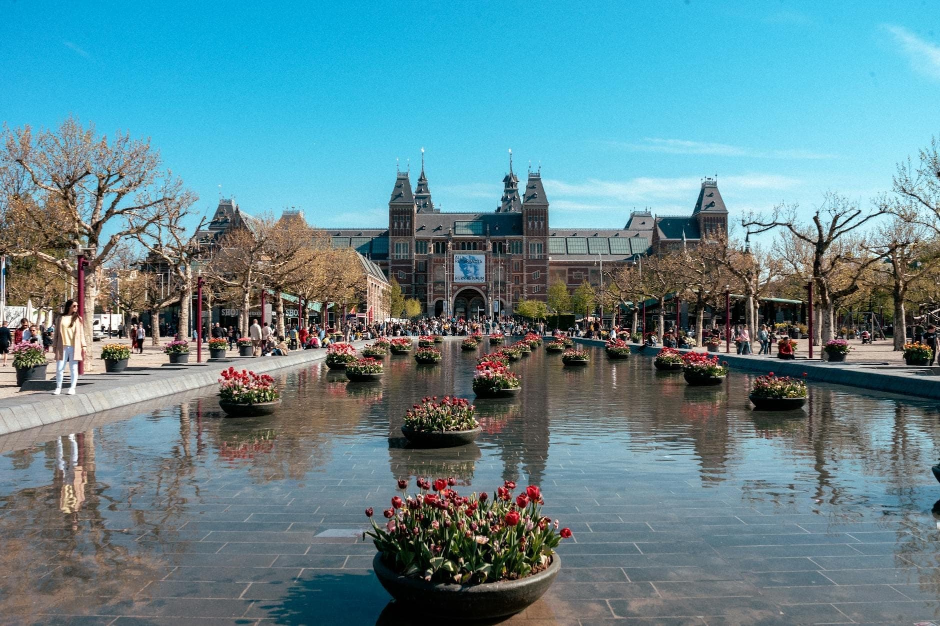 Weite Aufnahme des Rijksmuseums in Amsterdam mit großem Spiegelbecken und blühenden Blumen unter strahlend blauem Himmel, umgeben von Besuchern.