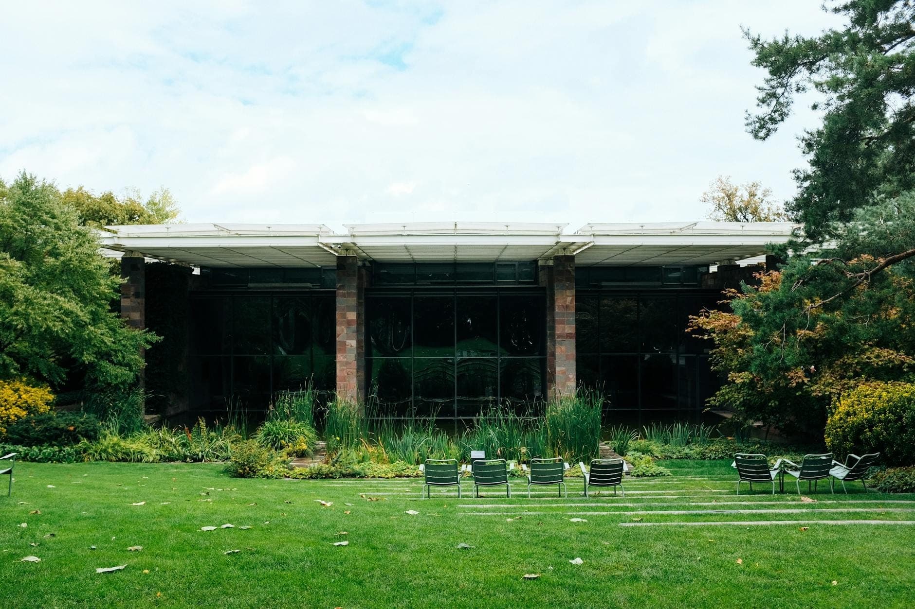 Pabellón modernista de cristal rodeado de césped verde y jardín paisajístico, con filas de sillas al aire libre y árboles a ambos lados.