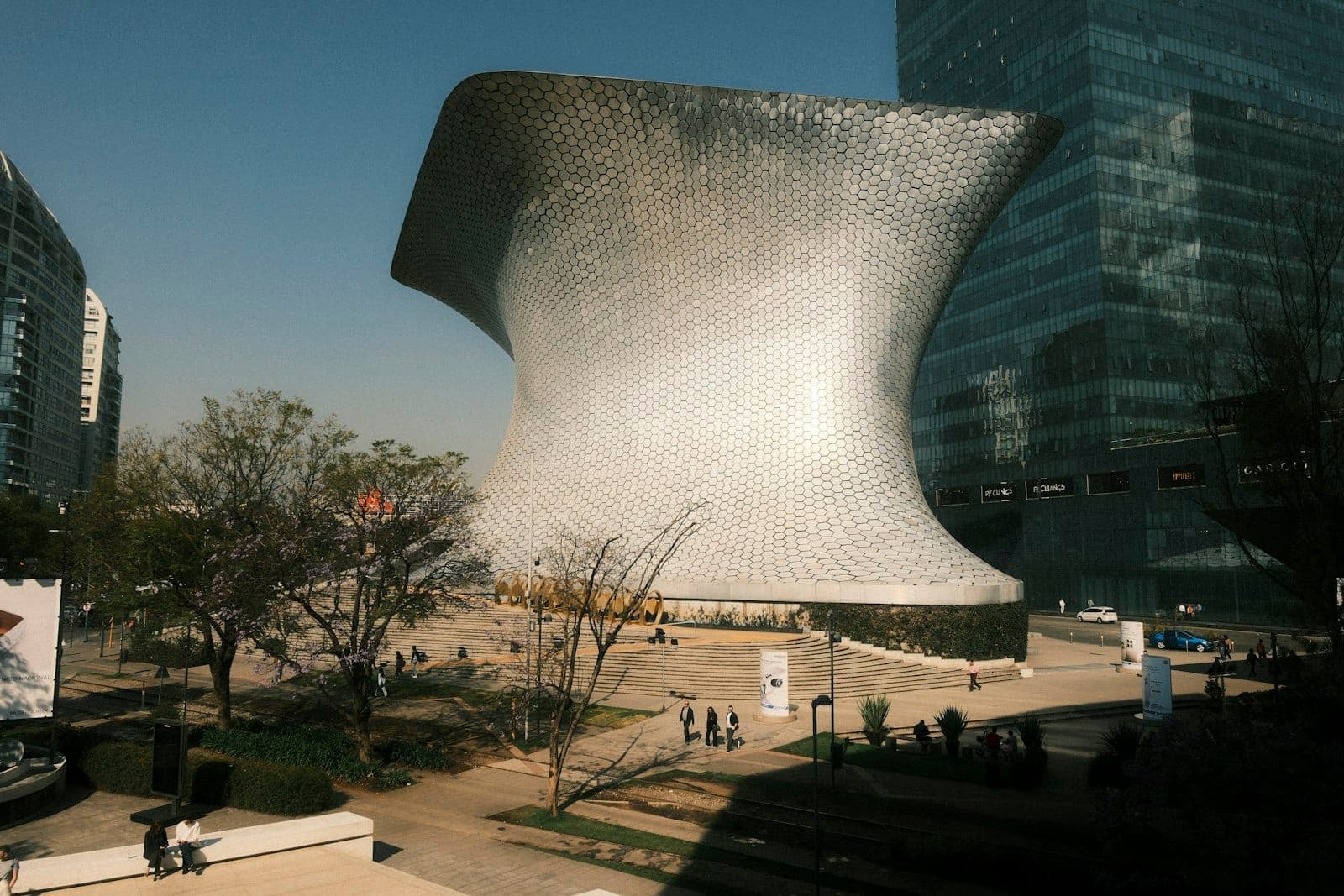 Striking silver hexagonal-tiled museum building with dramatic curves in a modern city plaza, people walking nearby in Polanco, Mexico City.
