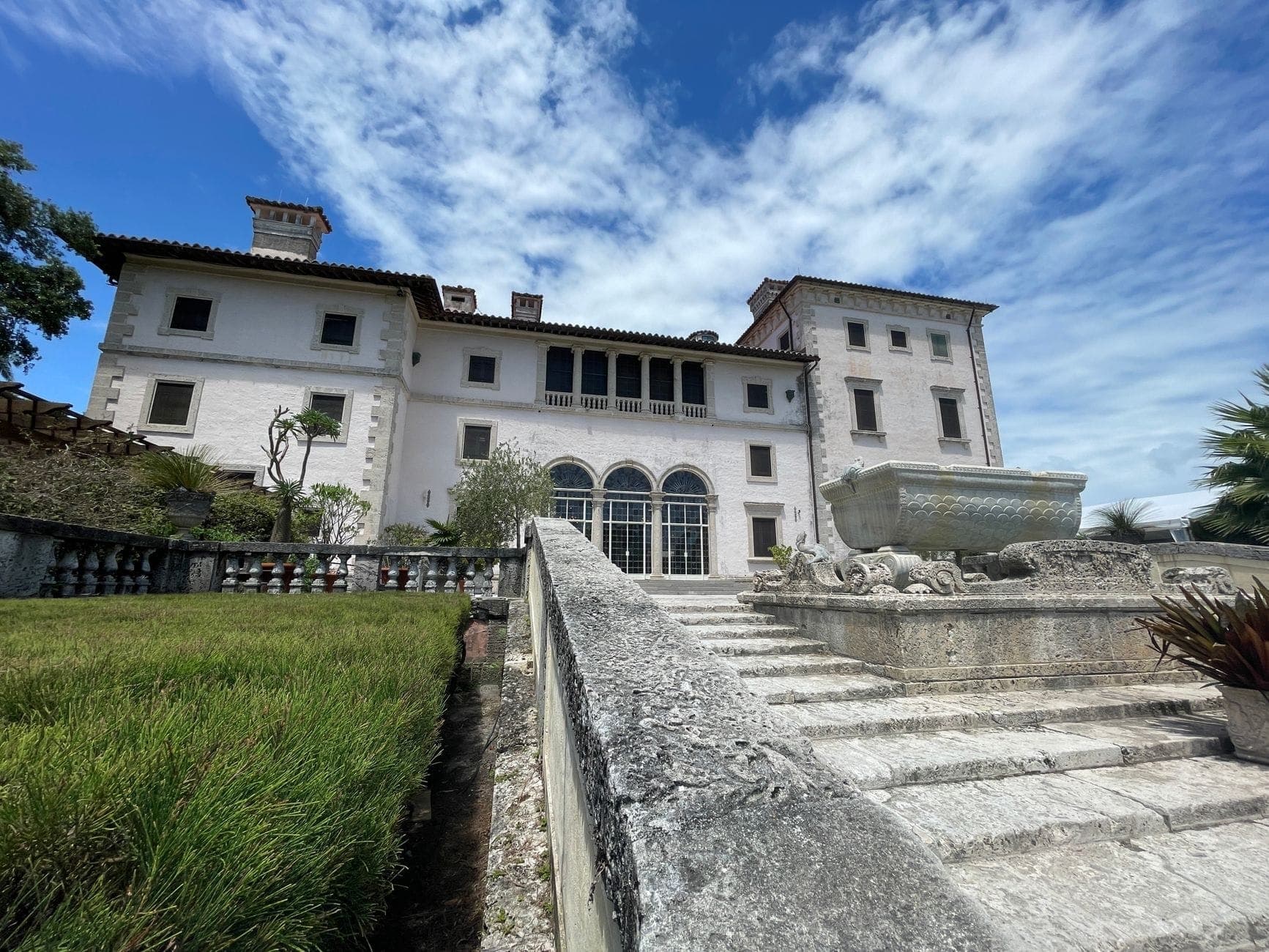 Historic mansion with Mediterranean architecture, stone steps, formal gardens and a large lawn under a blue sky with scattered clouds.