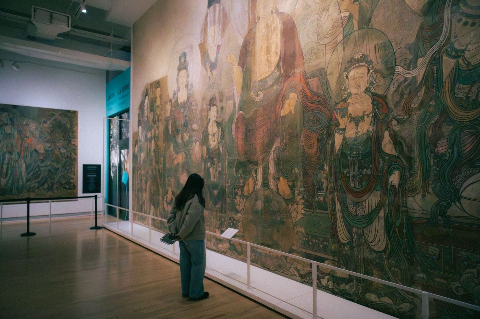 Seorang pengunjung mengagumi mural besar dan terperinci yang menampilkan tokoh-tokoh Asia tradisional di galeri yang terang benderang di sebuah museum di Toronto.