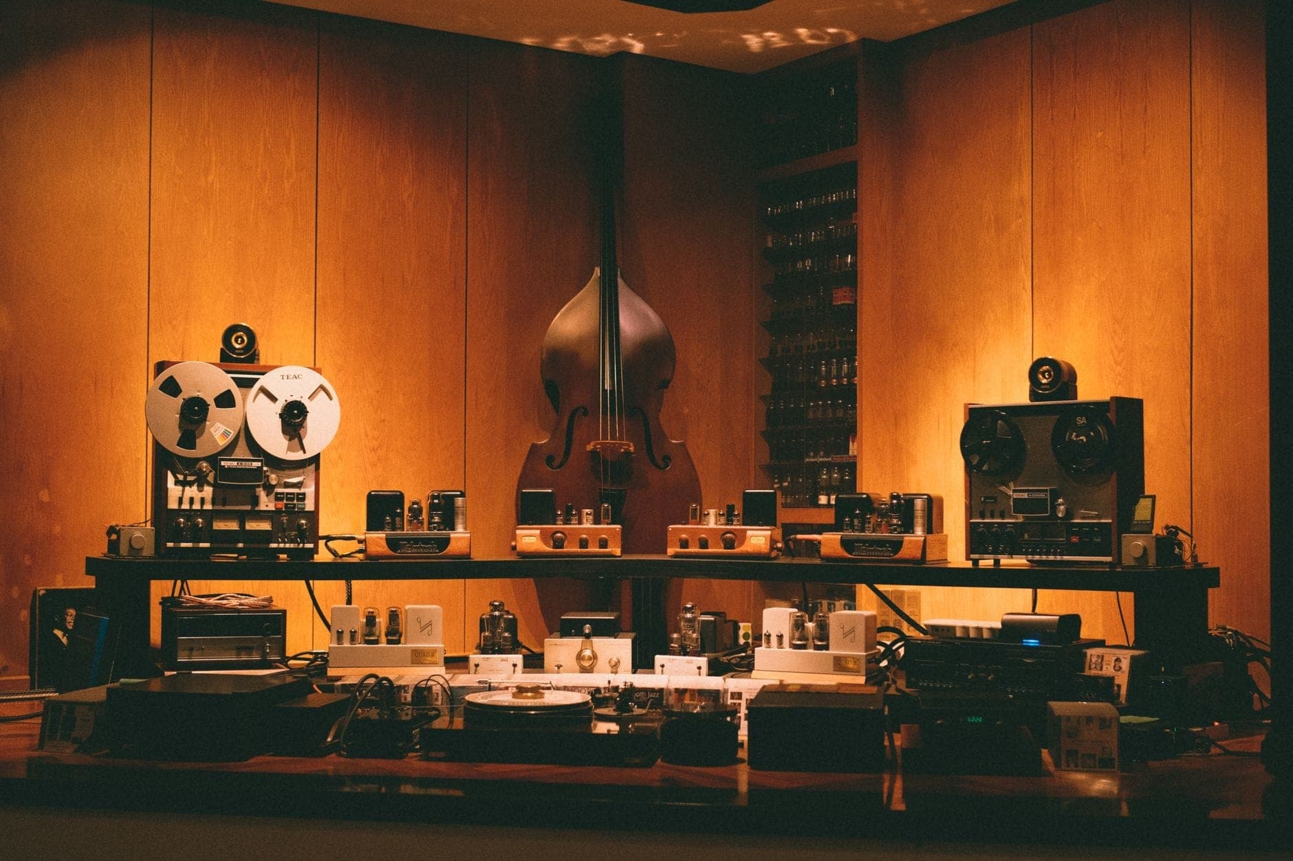 Warmbeleuchteter Musikstudioraum mit Tonbandmaschinen, Kontrabass und vintage Audiogeräten vor einer holzvertäfelten Wand.