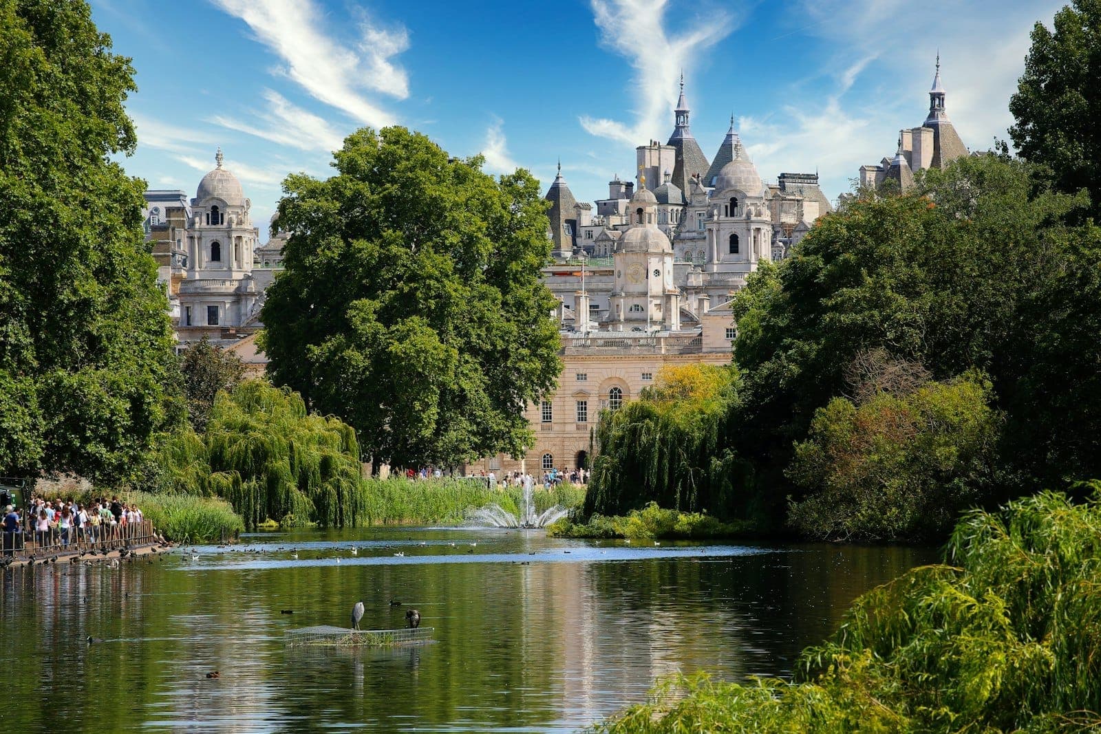 Blick über einen See in einem Londoner Park mit Enten, Grünflächen und imposanten historischen Gebäuden im Hintergrund.