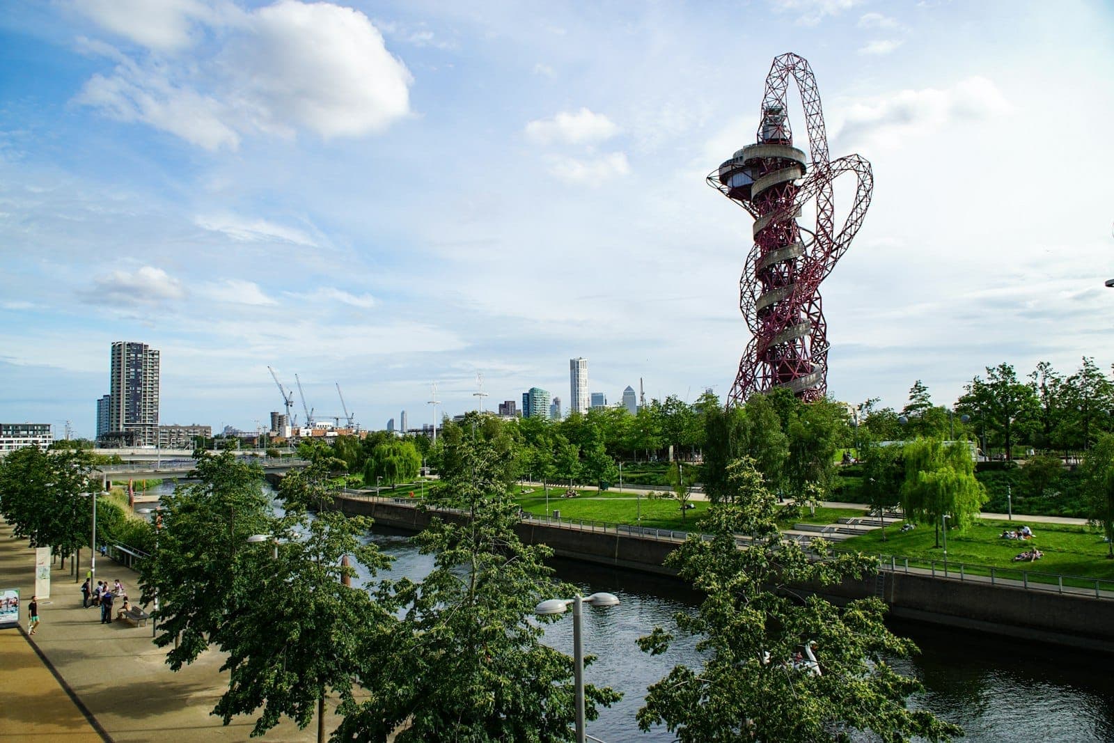 Blick auf den Queen Elizabeth Olympic Park in East London, mit der ArcelorMittal Orbit-Skulptur, Grünflächen, Bäumen, einem Fluss und Menschen, die sich im Freien entspannen.