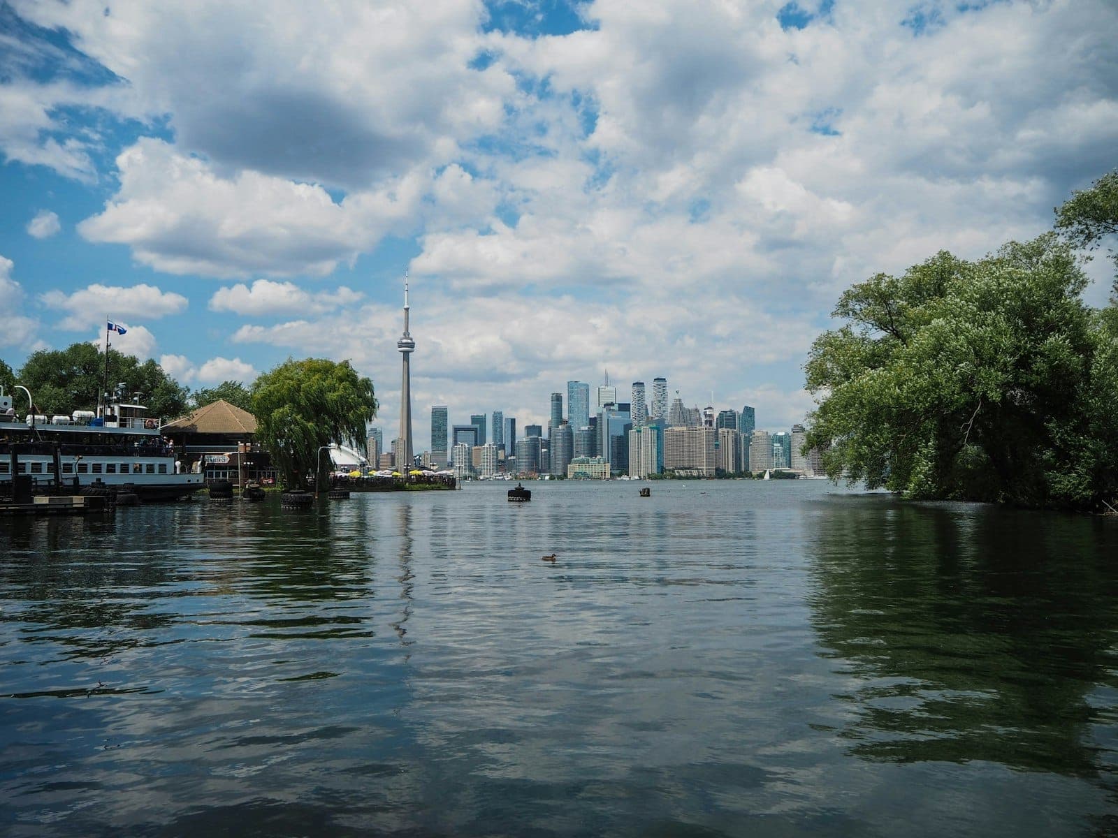 View of Toronto skyline from the Toronto Islands with water, lush greenery, and ferry boats in the foreground on a bright, partly cloudy day.