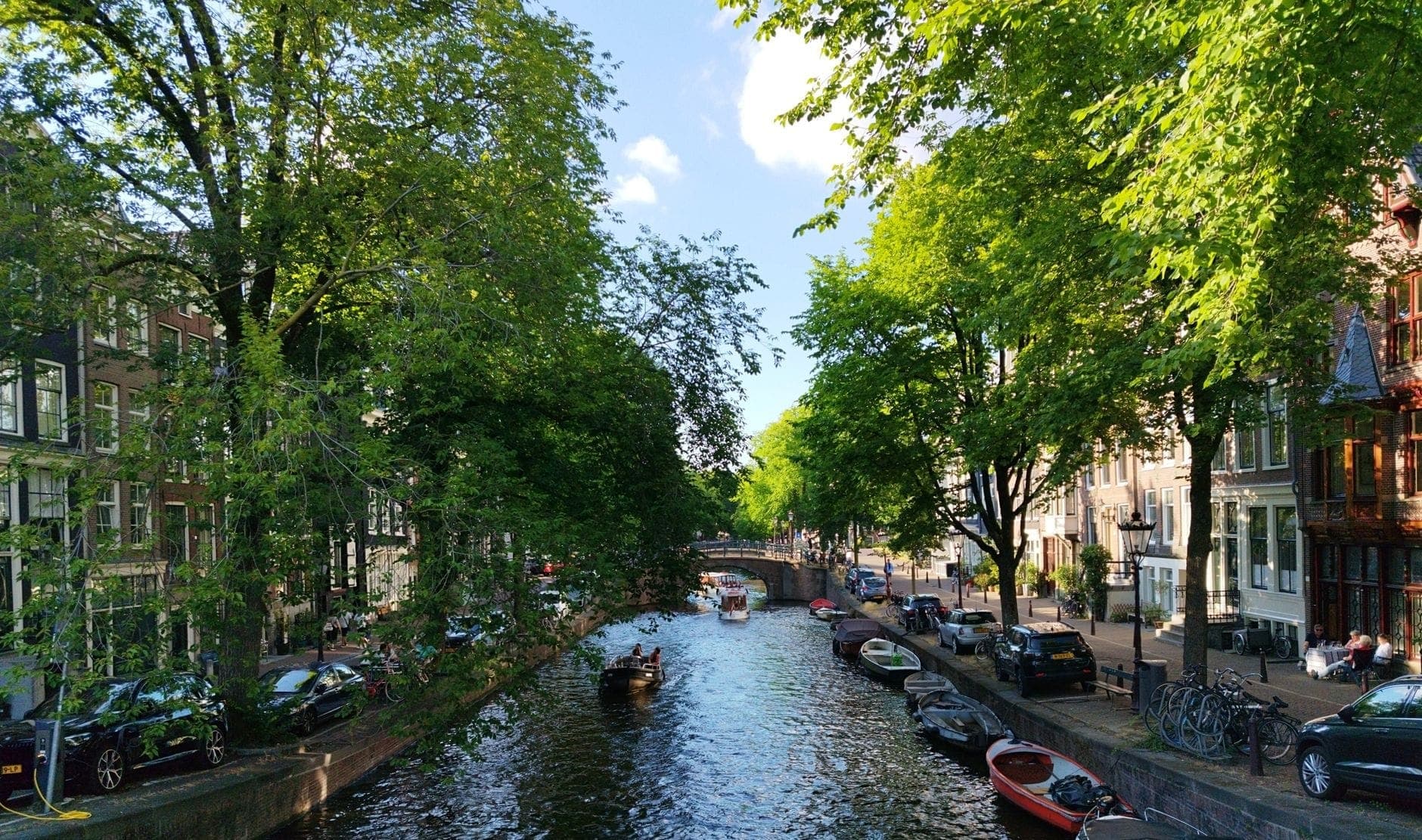 Sonniger Amsterdamer Kanal gesäumt von grünen Bäumen, Booten und typisch niederländischen Häusern – die lebendige Atmosphäre der Stadt bei schönem Wetter.