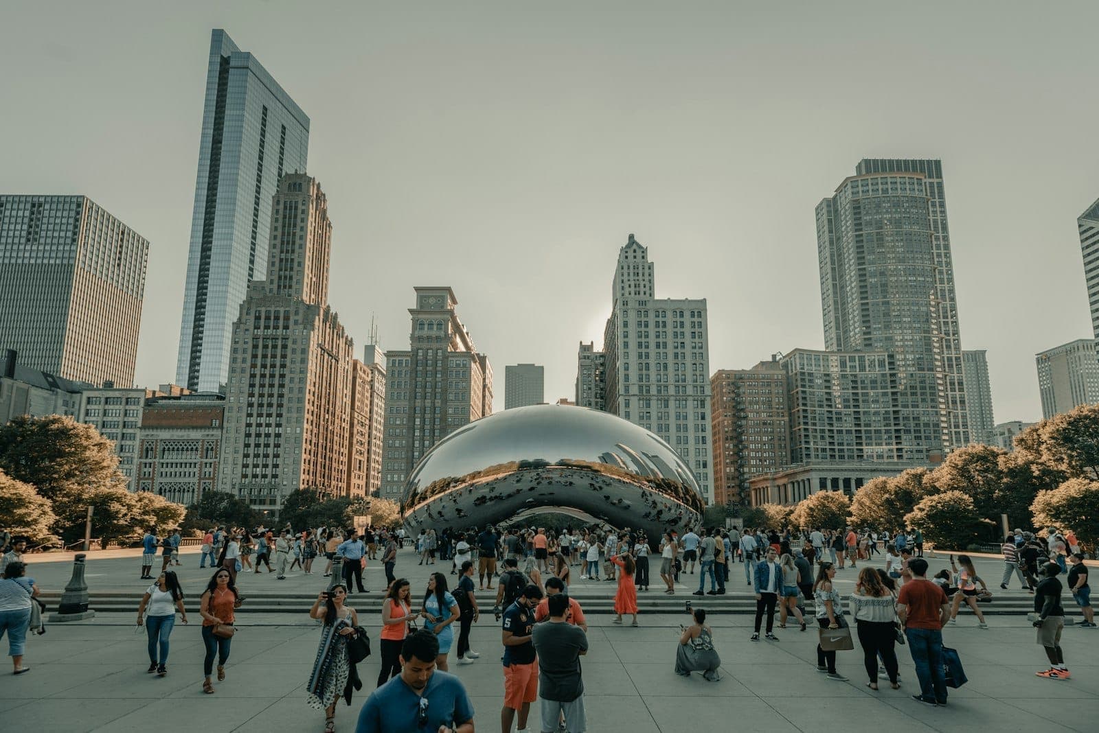 A lively crowd gathers around Chicago's iconic Cloud Gate sculpture in Millennium Park on a sunny day, with skyscrapers rising in the background.
