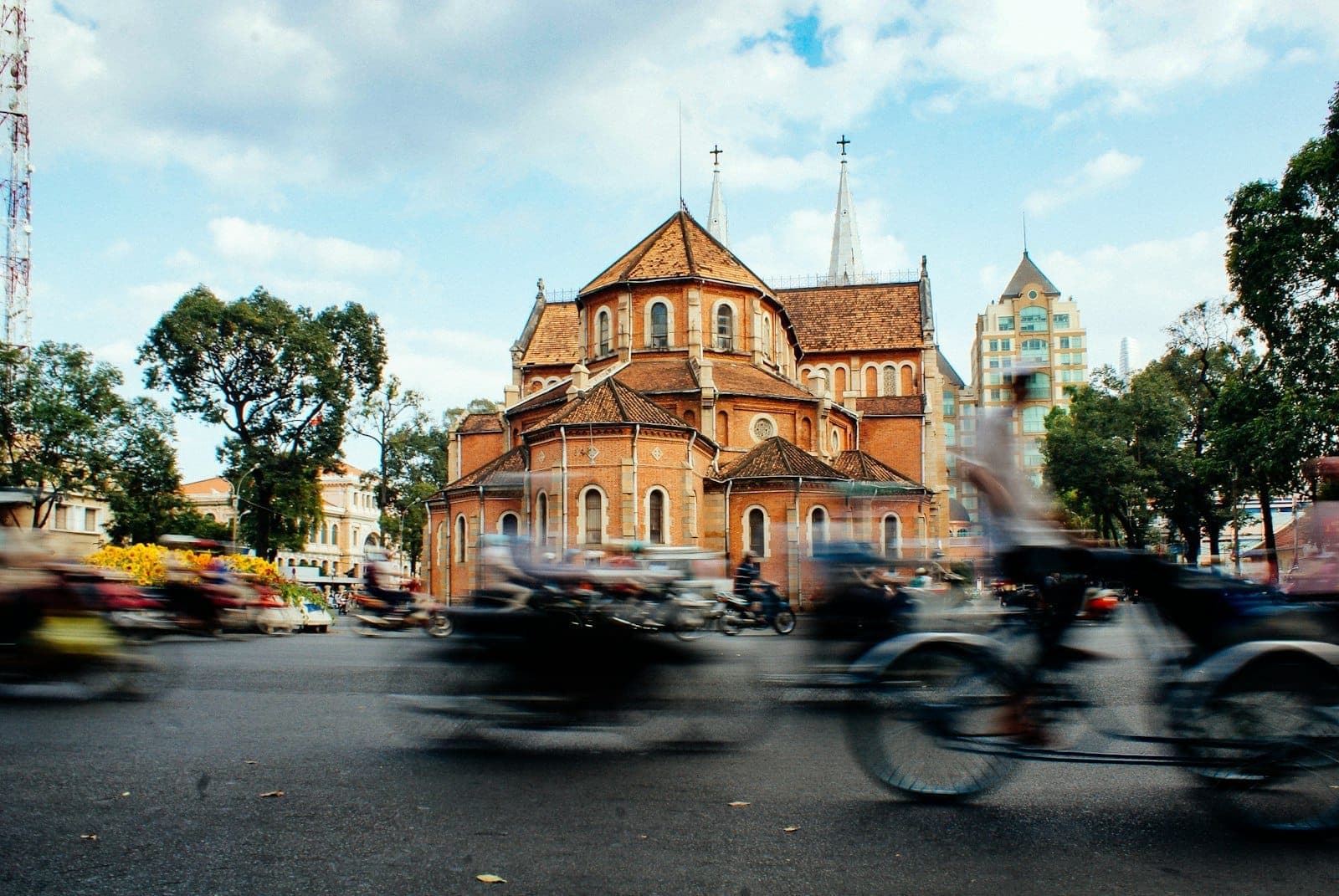 Motociclette che passano davanti alla Basilica Cattedrale di Notre-Dame di Saigon sotto un cielo azzurro con alcune nuvole a Ho Chi Minh City.