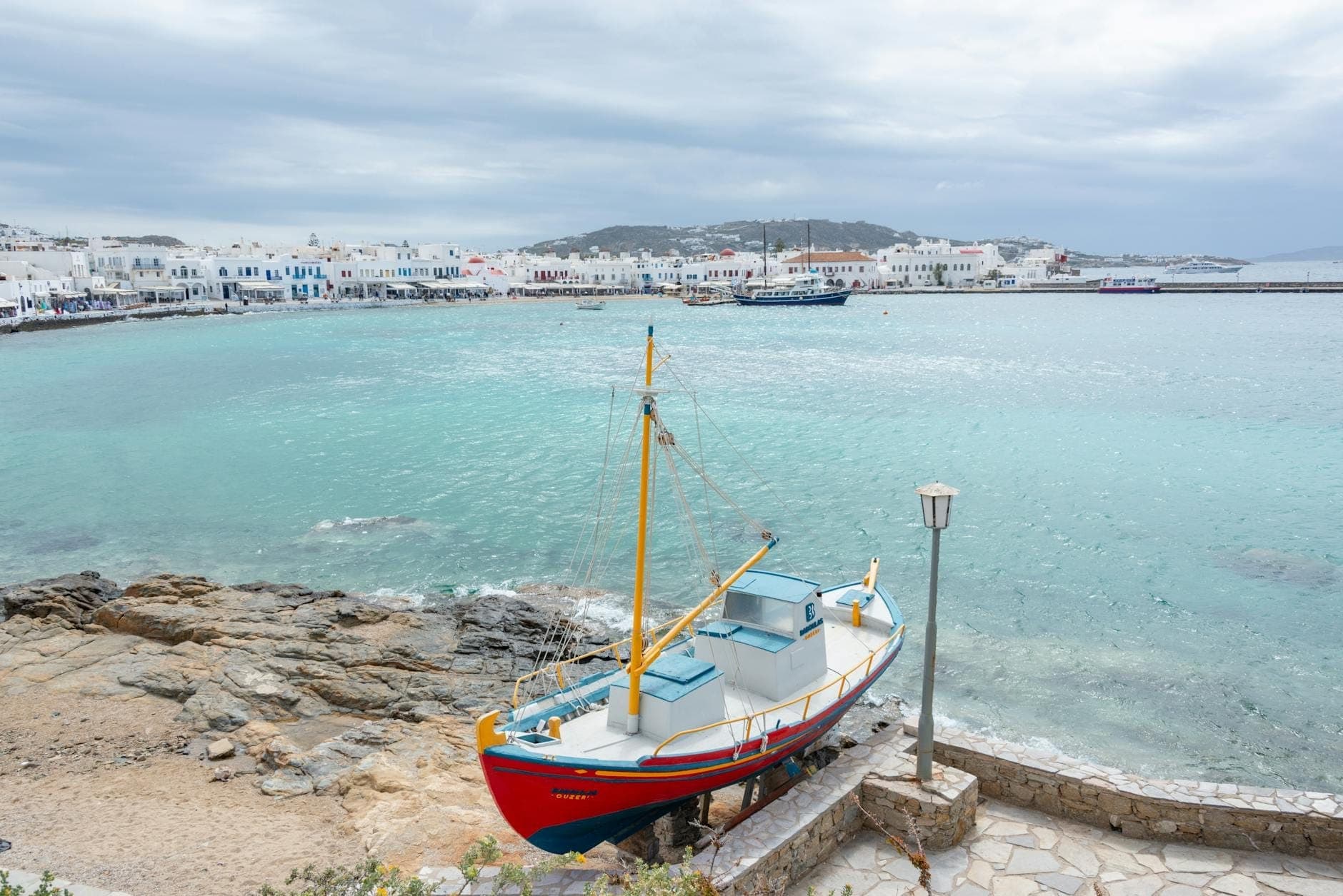 Ein gestrandetes Fischerboot an einem ruhigen, bewölkten Ufer mit der Stadt Mykonos und einer menschenleeren Küste im Hintergrund