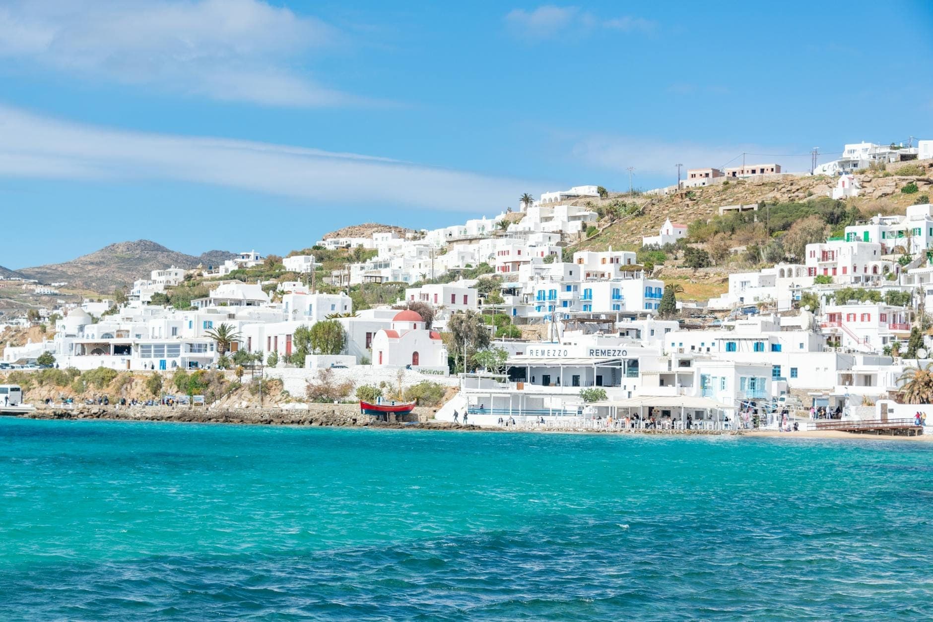 Hell erleuchtete Uferpromenade von Mykonos mit weiß getünchten Gebäuden auf einem Hügel und türkisfarbenem Meer unter blauem Himmel, als Illustration des mediterranen Klimas.