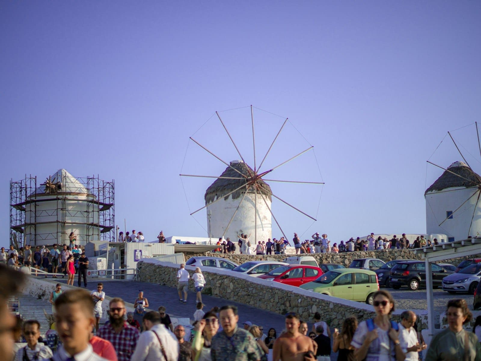 Belebte Szene an den Windmühlen von Mykonos mit vielen Touristen und Autos unter einem klaren Himmel, die das Gedränge in der Hochsaison veranschaulicht.