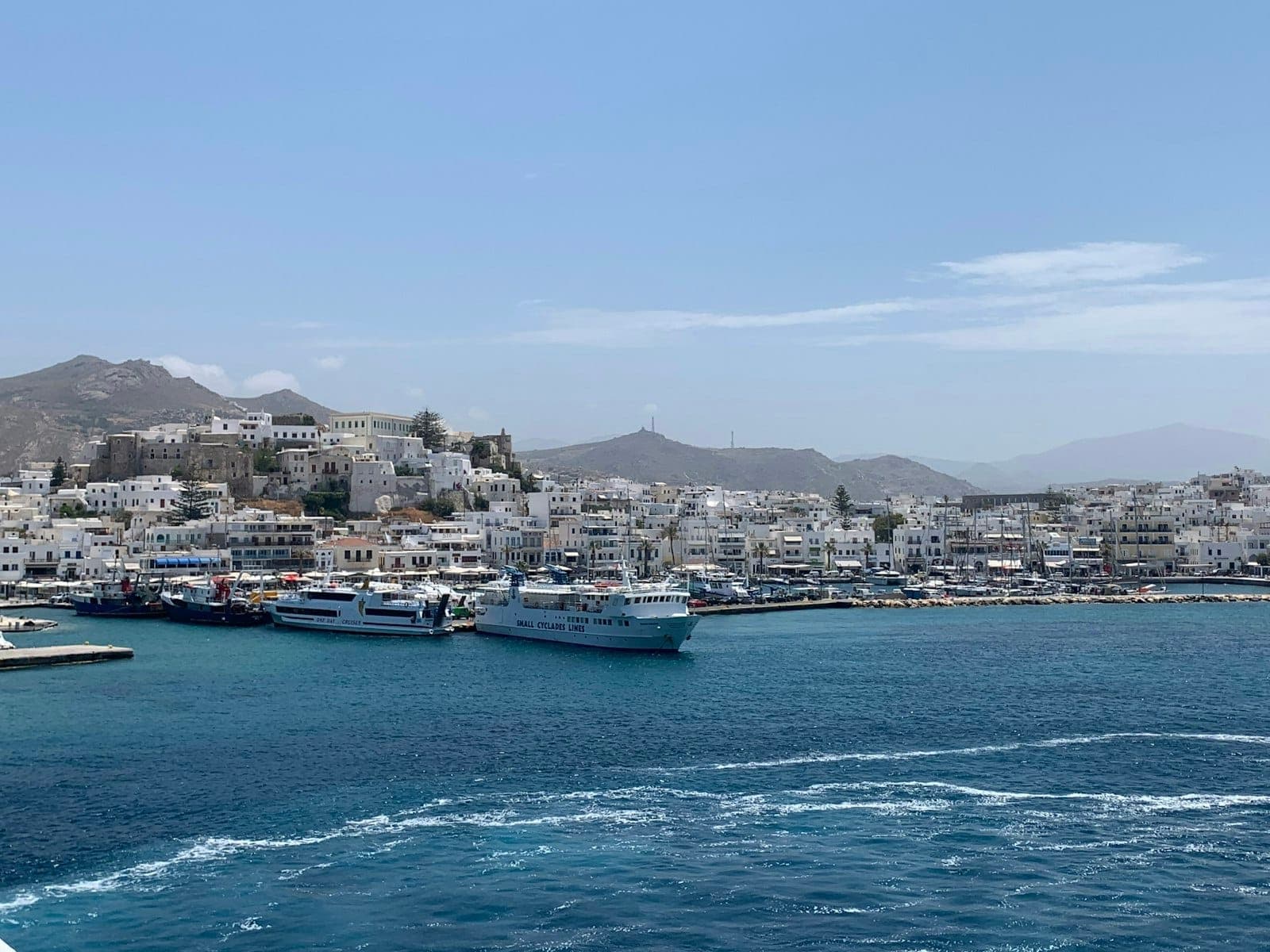 Fähren und Boote im Hafen von Mykonos mit weiß getünchten Stadtgebäuden im Hintergrund unter einem klaren Himmel.