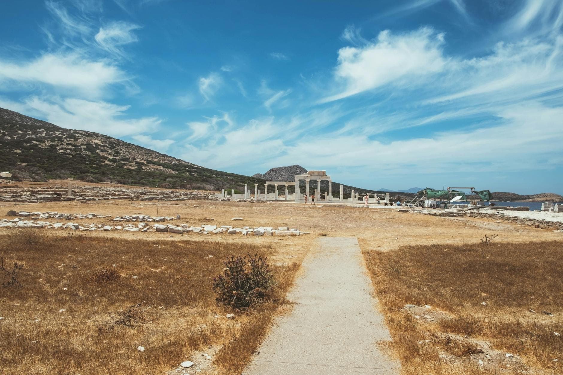 Weg zu antiken Ruinen unter blauem Himmel, mit Säulen und Hügeln im Hintergrund auf Mykonos.