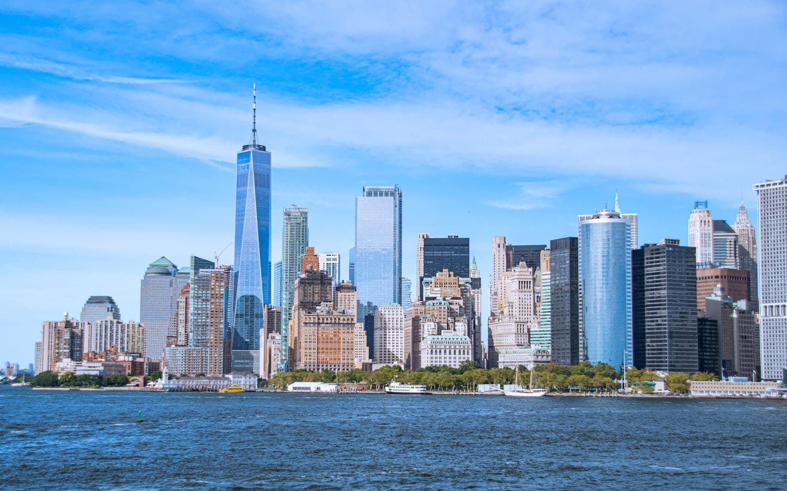 วิวกลางวันของเส้นขอบฟ้า Lower Manhattan ในนิวยอร์กซิตี้ ท้องฟ้าสีฟ้าใส และแม่น้ำ Hudson อยู่เบื้องหน้า