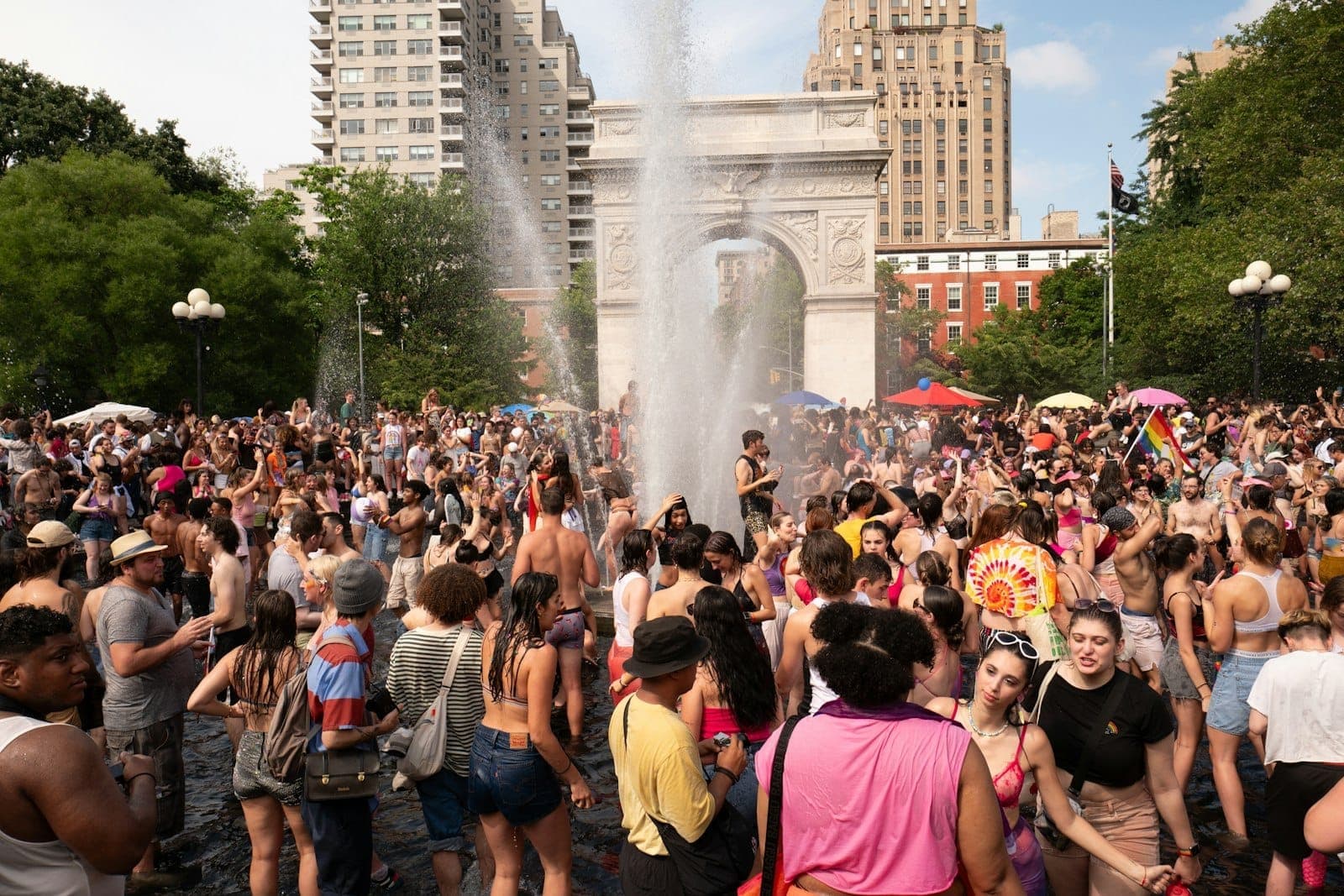 ฝูงชนจำนวนมากสนุกสนานในวันฤดูร้อนรอบๆ น้ำพุที่ Washington Square Park โดยมีซุ้มประตูและเส้นขอบฟ้าของเมืองเป็นฉากหลัง