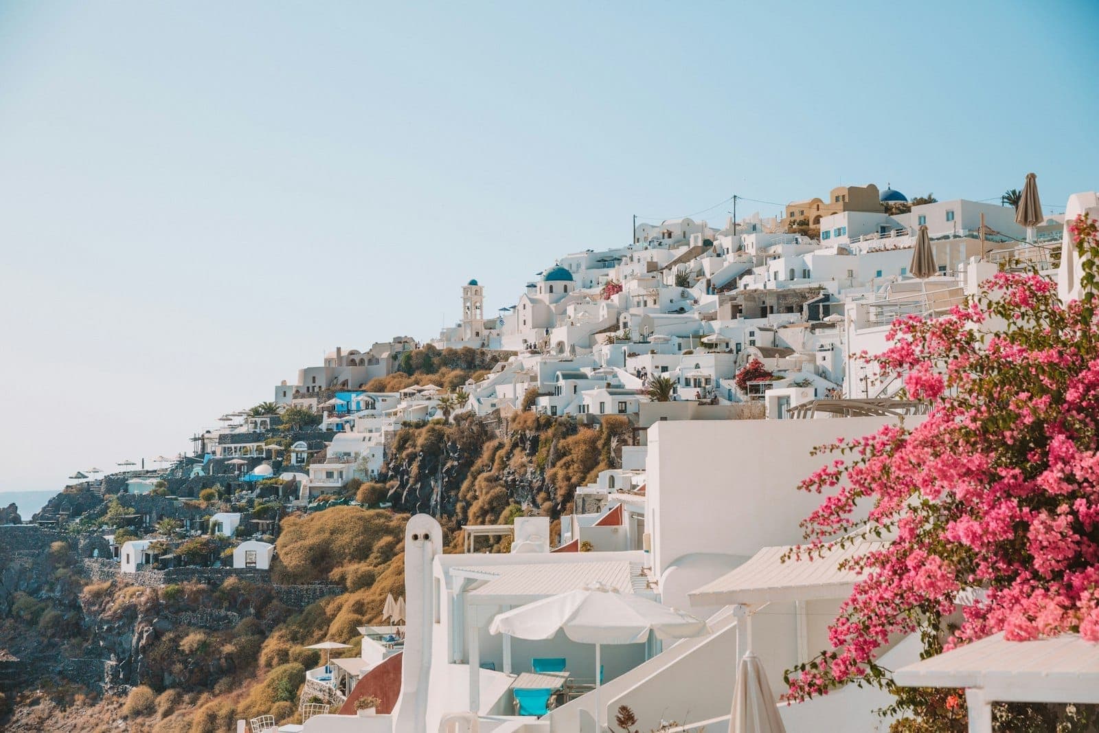 Vista clássica do penhasco de Santorini com edifícios caiados de branco, cúpulas azuis e buganvílias cor-de-rosa sob um céu azul limpo, transmitindo uma atmosfera calma e agradável.