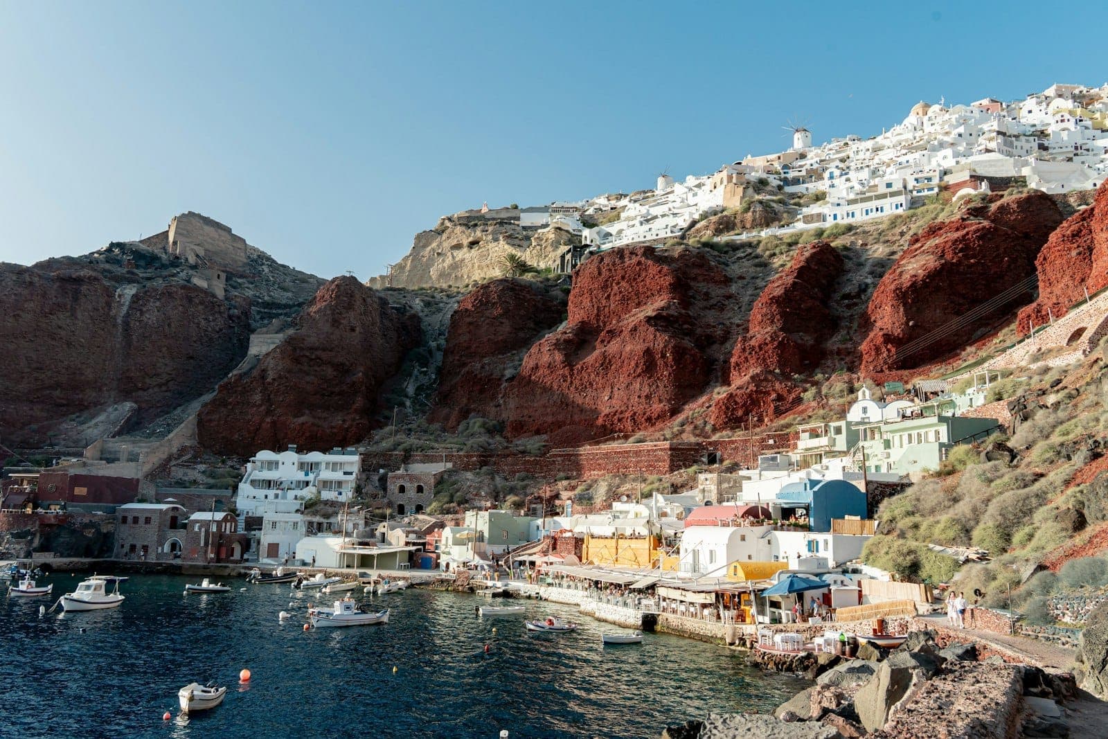 Porto de Santorini com restaurantes tradicionais à beira-mar, barcos na água e edifícios caiados na encosta, ilustrando a atividade sazonal e os negócios locais.