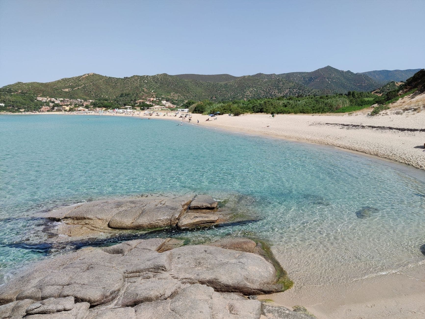 Ruhiger Sandstrand mit türkisfarbenem Wasser, einige Personen in der Ferne, Hügel und Grün unter klarem Himmel, typisch für die Nebensaison auf Sardinien