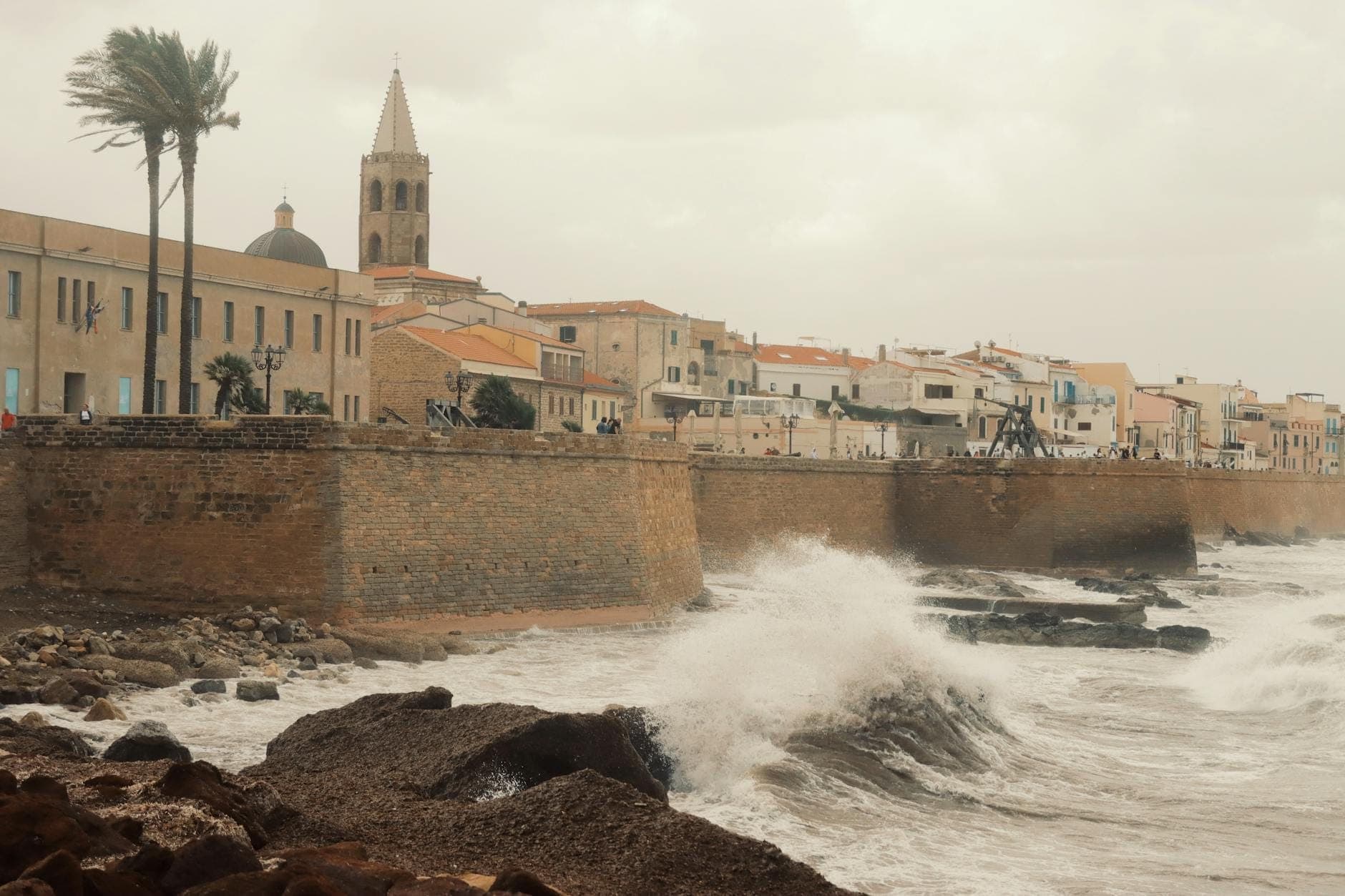 Wellen brechen gegen Felsen und eine historische Steinmauer unter einem bewölkten Himmel in einer Küstenstadt Sardiniens und erinnern an Winterwetter.