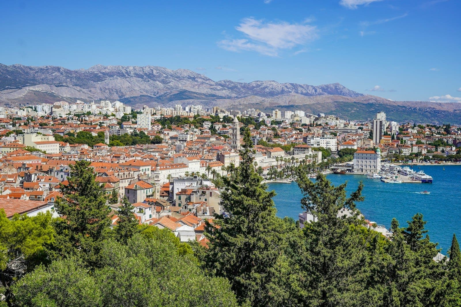 Vista panoramica di Spalato, Croazia, con la città vecchia storica, il porto fiancheggiato da palme, il Mare Adriatico azzurro e le montagne sullo sfondo sotto un cielo luminoso.
